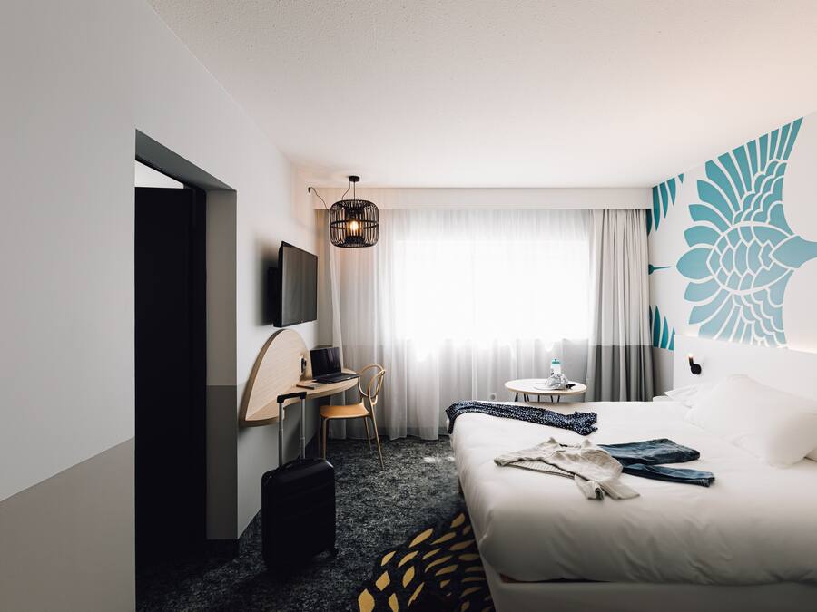 A hotel room with a bed, a desk, a chair, a suitcase, a TV, and a window with curtains.