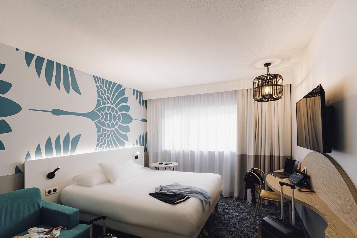 Une chambre d’hôtel comprenant un lit, un bureau, une chaise, un téléviseur et un mur orné d’un motif de feuilles.