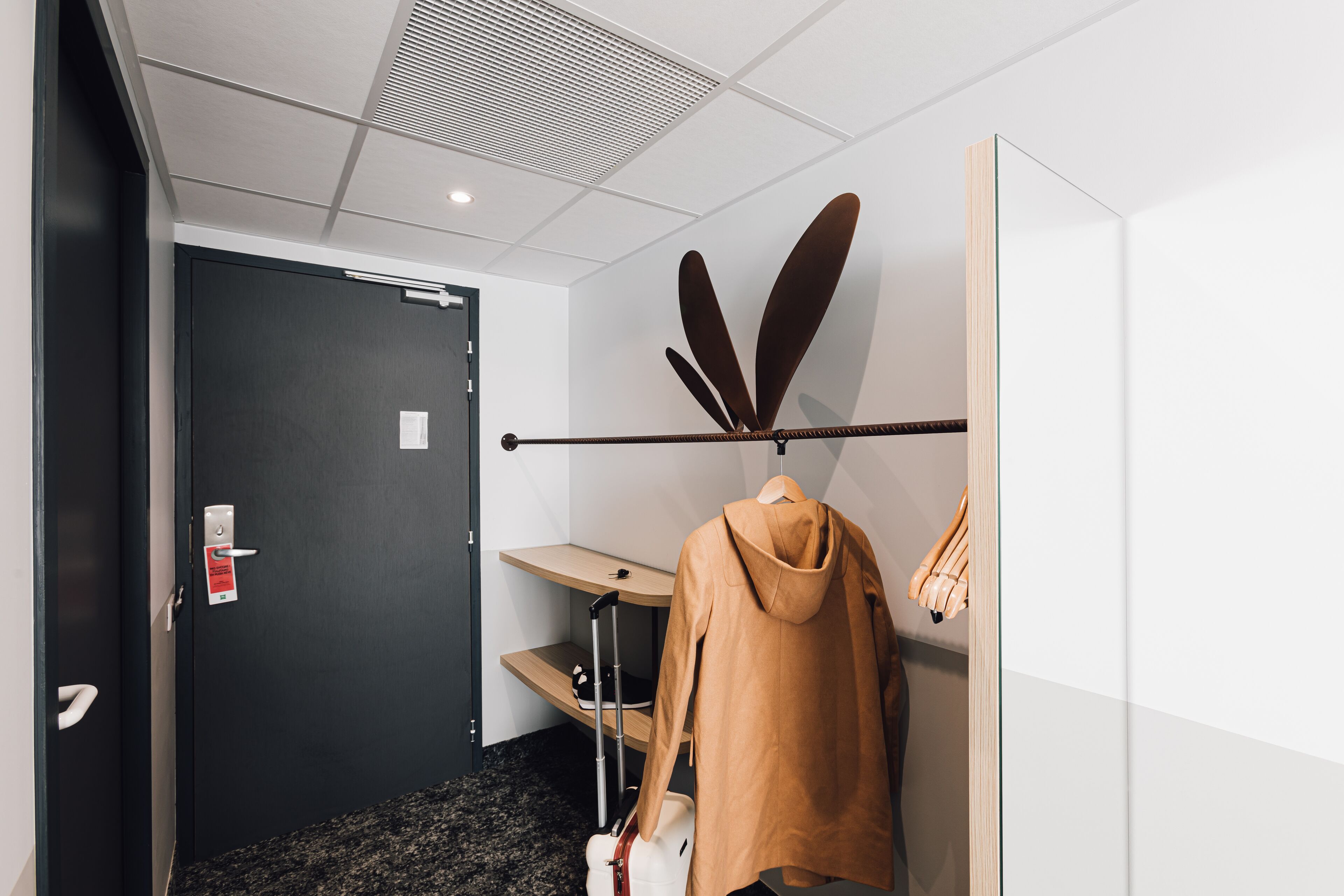 A hotel room with a dark door, a coat rack with a brown jacket, a small table, and a suitcase.