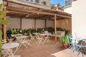 An outdoor patio with white metal tables and chairs, surrounded by potted plants and a wooden pergola. - Parker (Rome)
