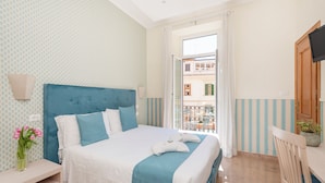 A hotel room with a bed, a nightstand, a chair, a TV, and a balcony with a view of buildings.