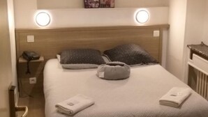 A neatly made bed with a patterned bedspread, two pillows, and a bedside lamp. There is a small bedside table with a lamp, a wall clock, and a framed picture above the bed. The room has a wooden floor and a ceiling light fixture. - Hôtel Pacific (Calais)