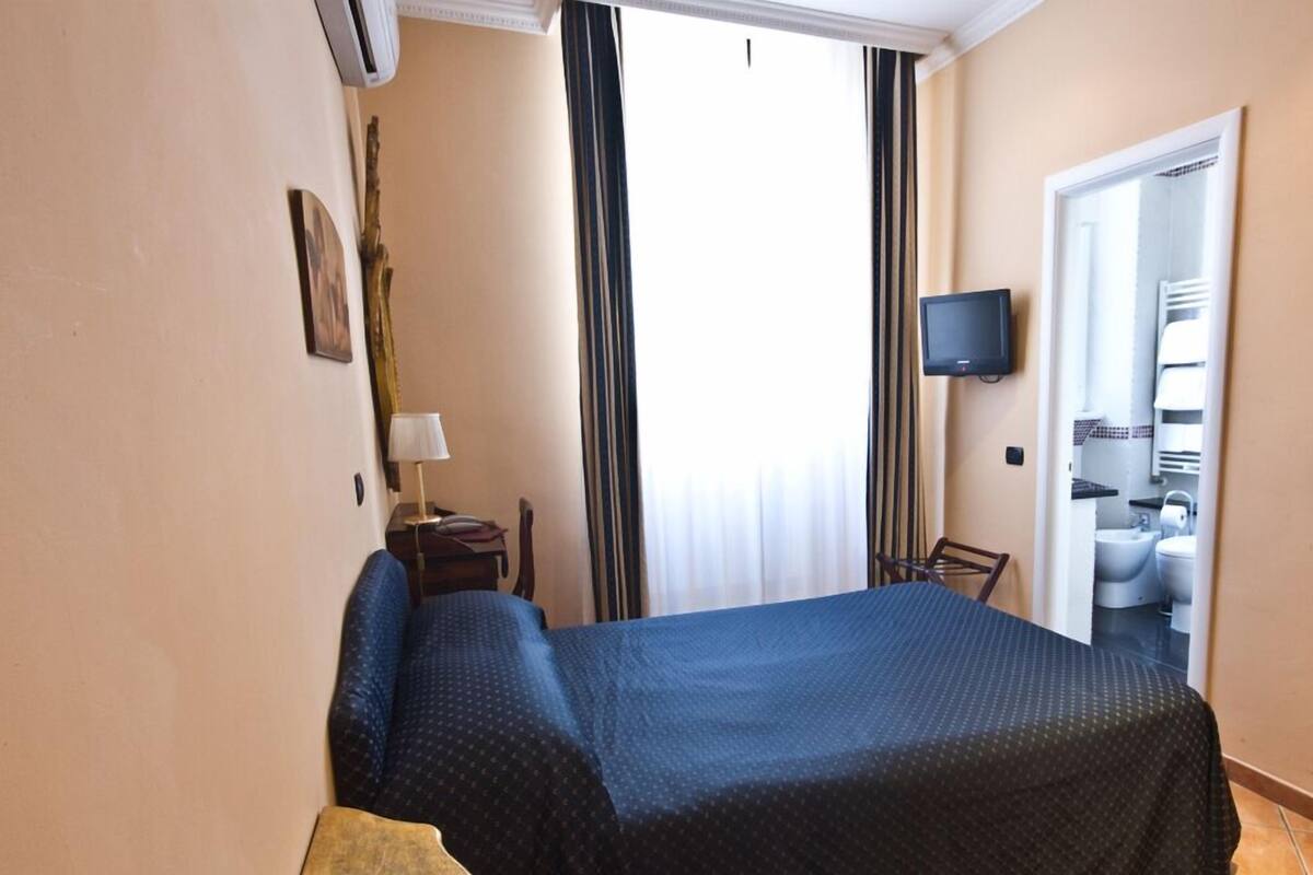 A hotel room with a bed, a desk with a lamp, a TV mounted on the wall, and a bathroom visible through an open door.