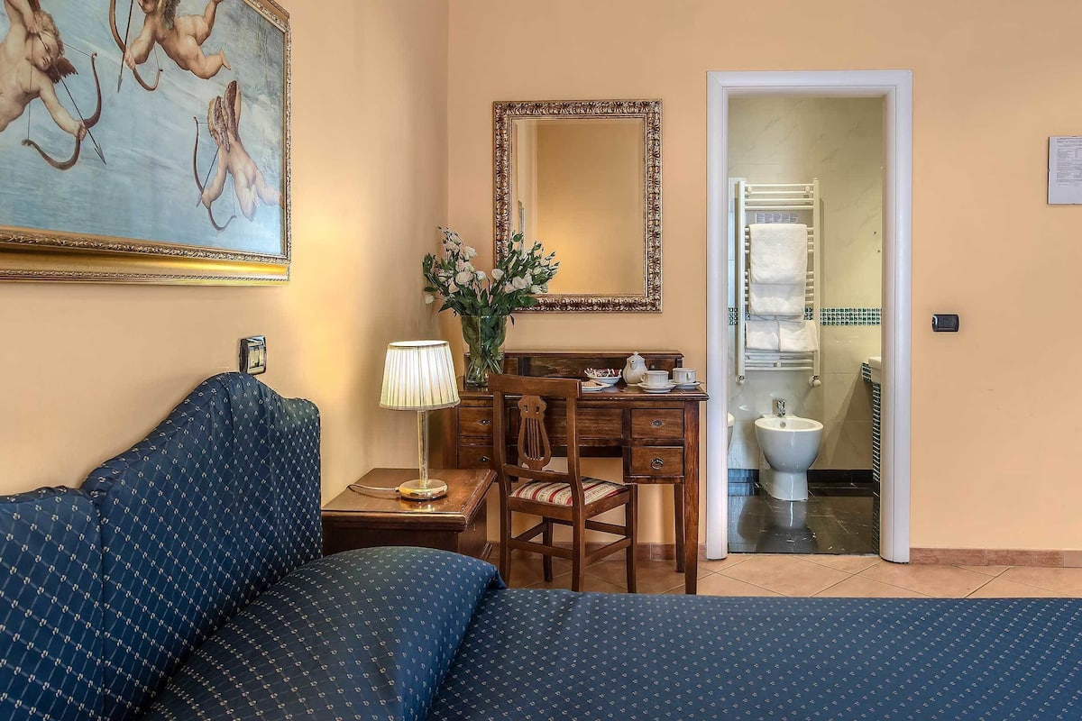 A hotel room with a bed, a desk, a chair, a mirror, and a bathroom visible through an open door.
