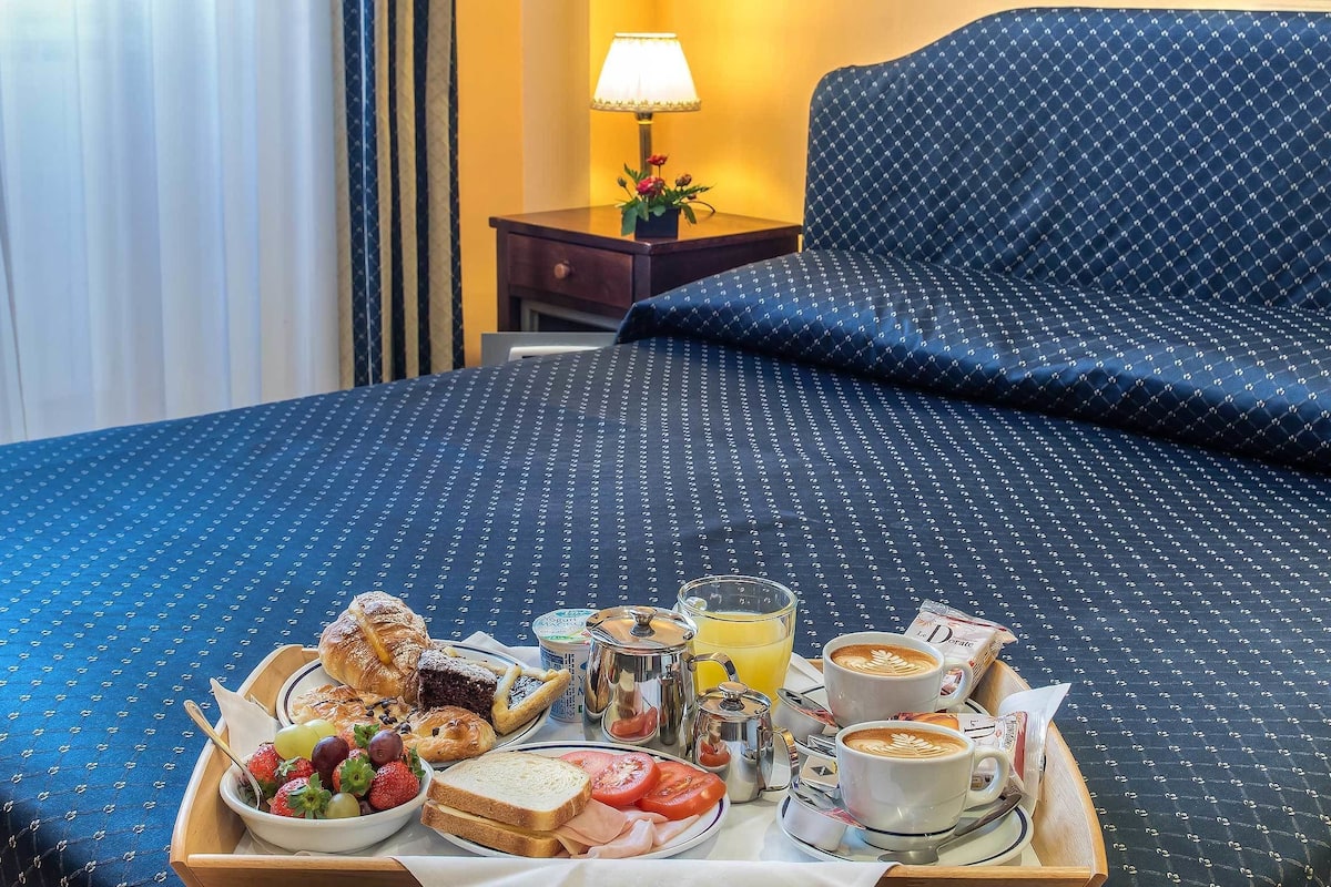 A neatly made breakfast tray on a bed with a blue patterned bedspread, a nightstand with a lamp and flowers, and a view of a hotel room.