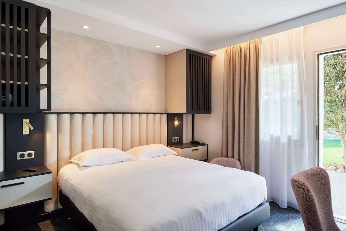 Una habitación de hotel moderna con una cama grande, una mesita de noche con lámpara, un televisor montado en la pared y vistas al exterior a través de una ventana.