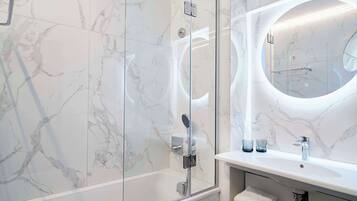 Een moderne badkamer met een douche met glazen wand, een wand betegeld met marmer en een ronde spiegel boven een wit wastafel.
