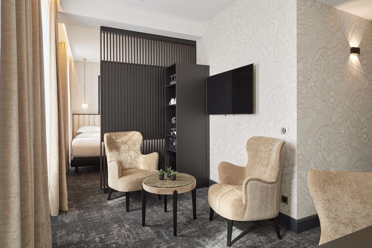 Una habitación de hotel moderna con una cama, dos sillones, una mesita redonda, una estantería y un televisor de pantalla plana montado en la pared.