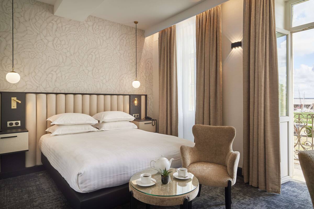 Una habitación de hotel con una cama grande, una silla, una mesita de vidrio con una tetera y tazas, y cortinas.