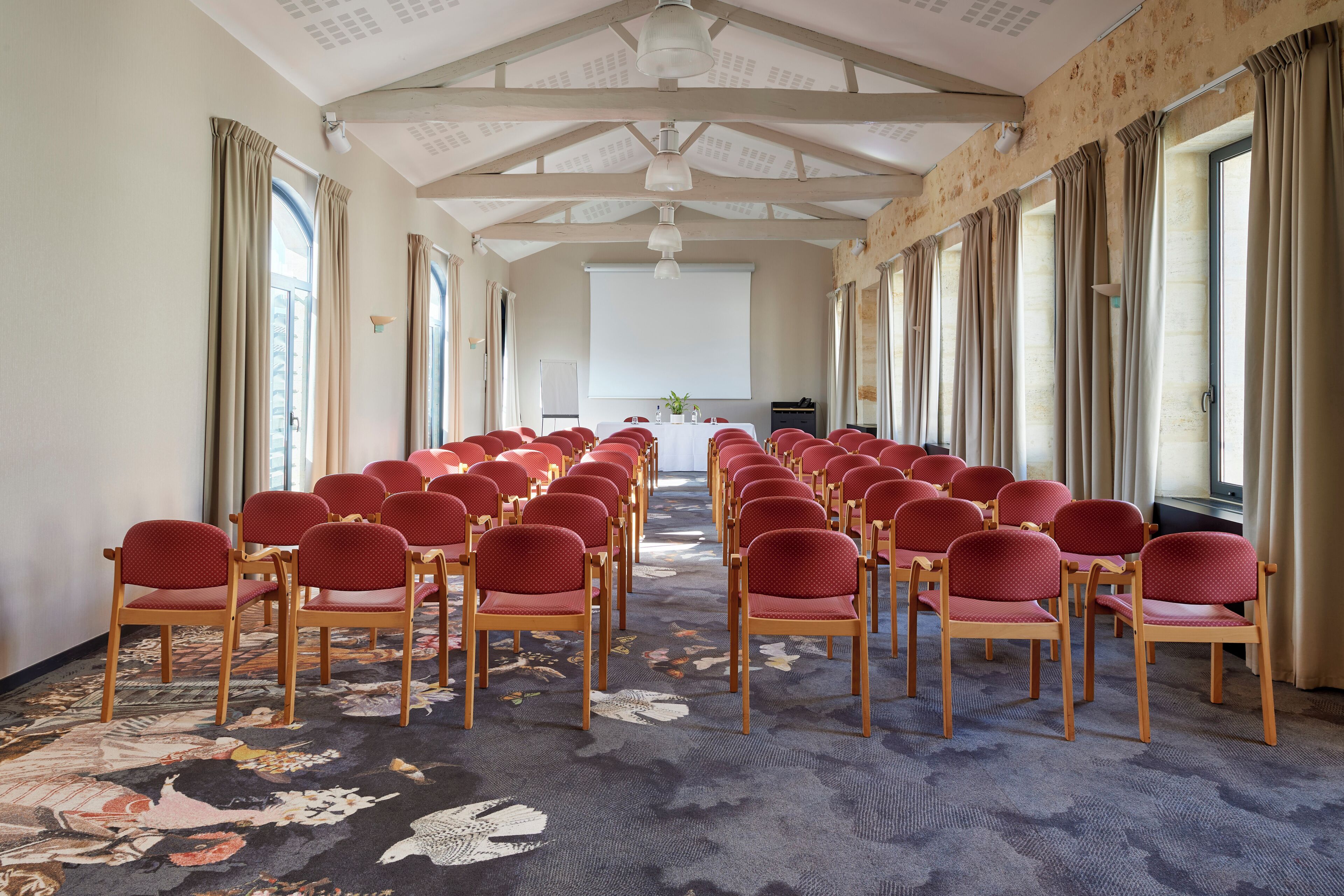 Un salón de conferencias con hileras de sillas rojas, piso alfombrado y ventanas amplias con cortinas.