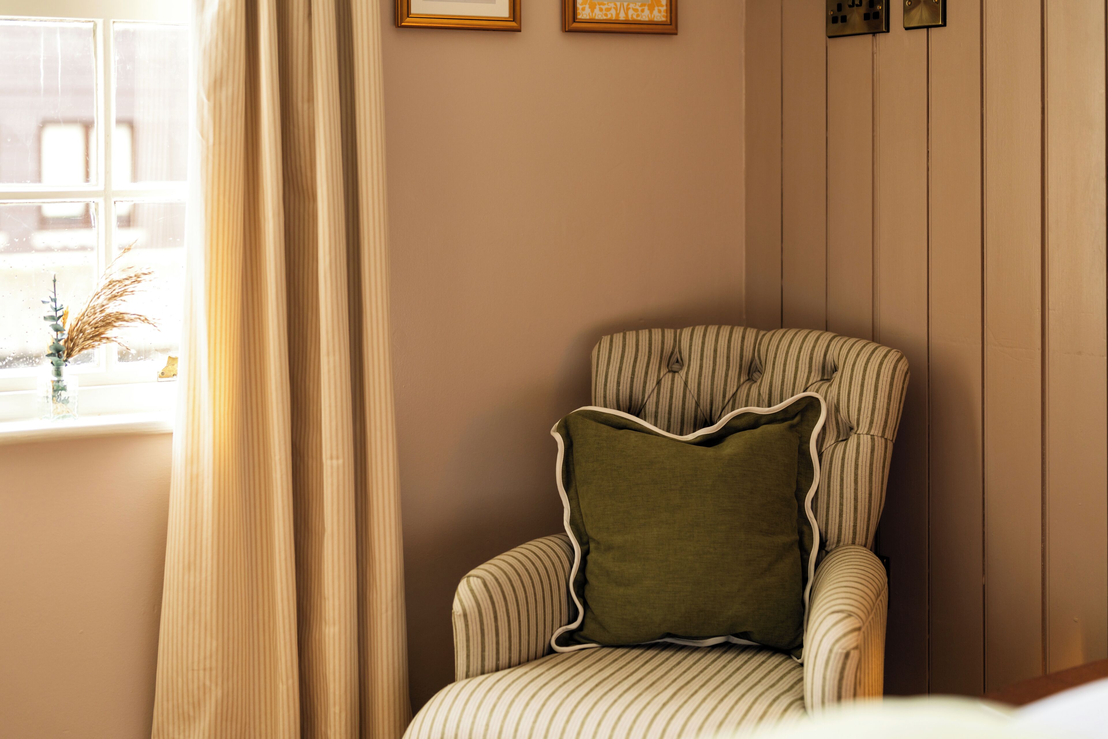 Une pièce d’angle avec un fauteuil à rayures, une fenêtre avec des rideaux et des tableaux encadrés accrochés au mur.