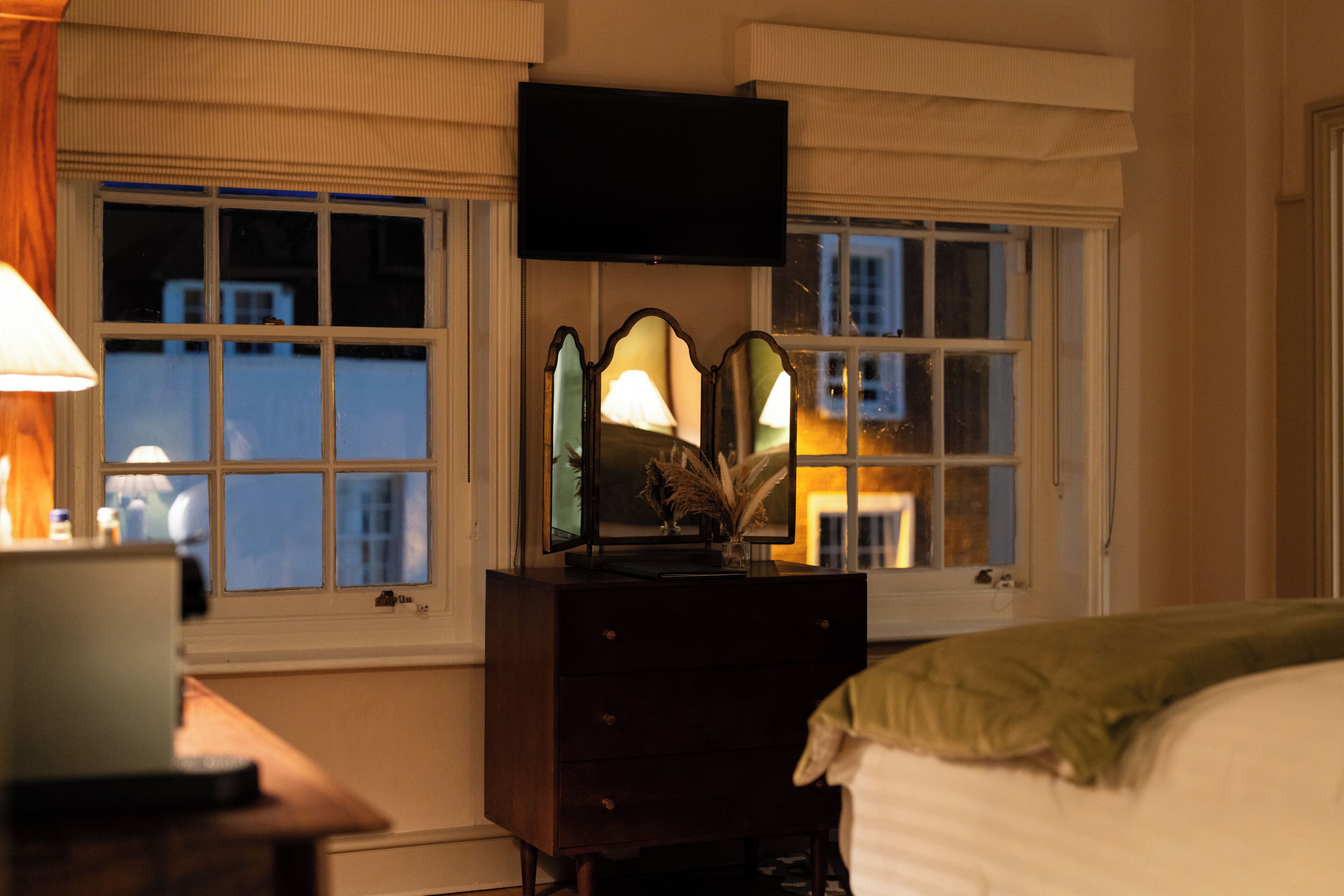 Un dormitorio con cama, cómoda, lámpara y televisor sobre una ventana.