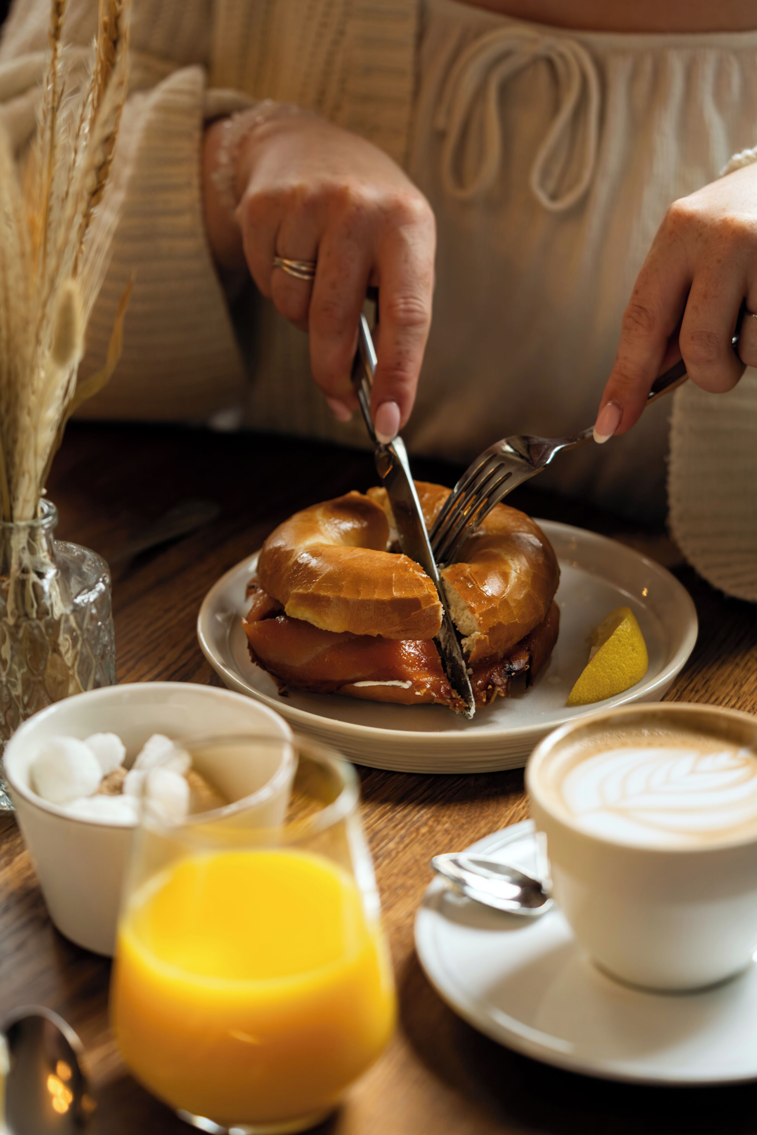 Een persoon snijdt een sandwich op een bord, met een partje citroen, een glas jus d'orange en een kop koffie op een houten tafel.