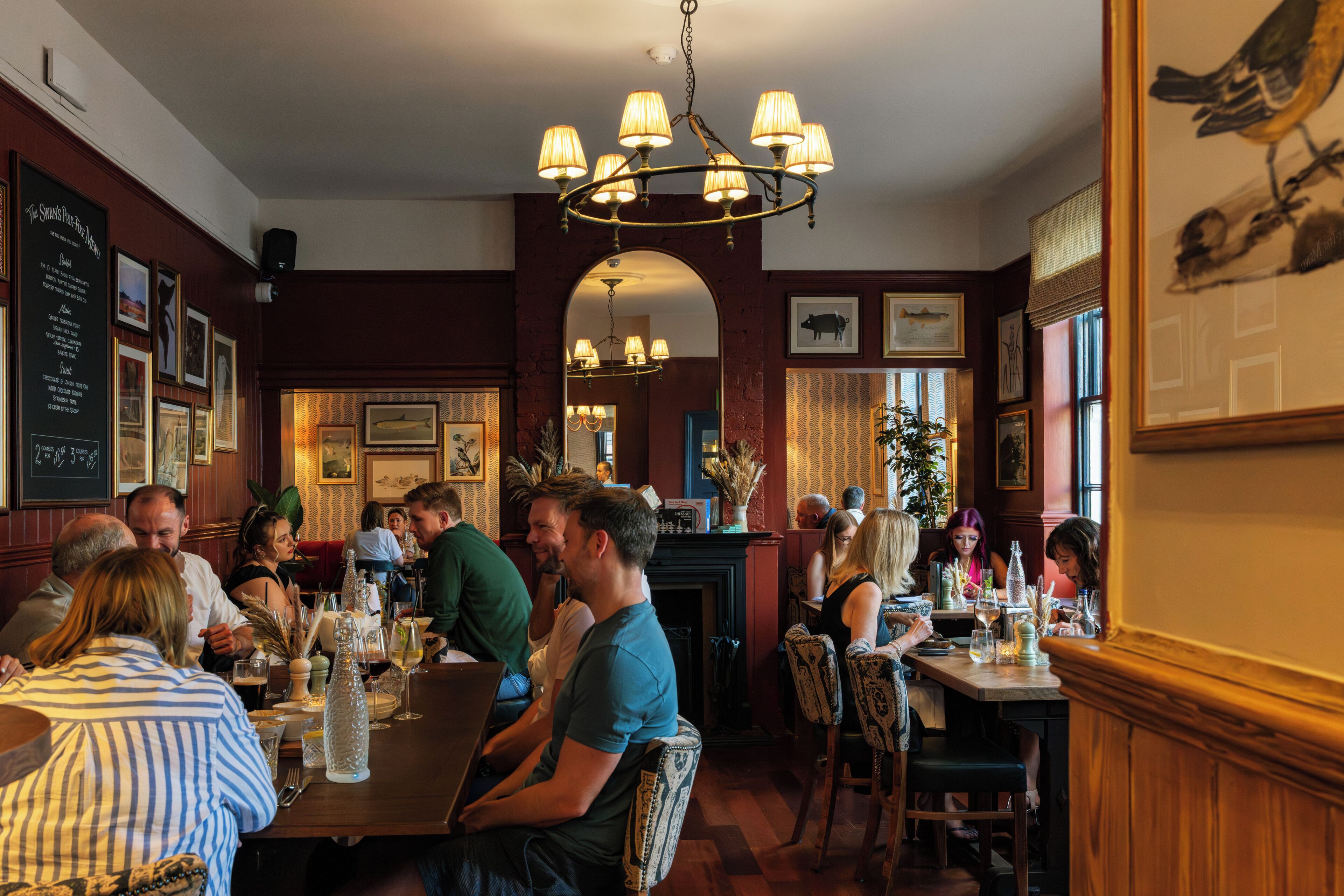 A restaurant interior with wooden paneling, chandeliers, framed artwork, and patrons dining.