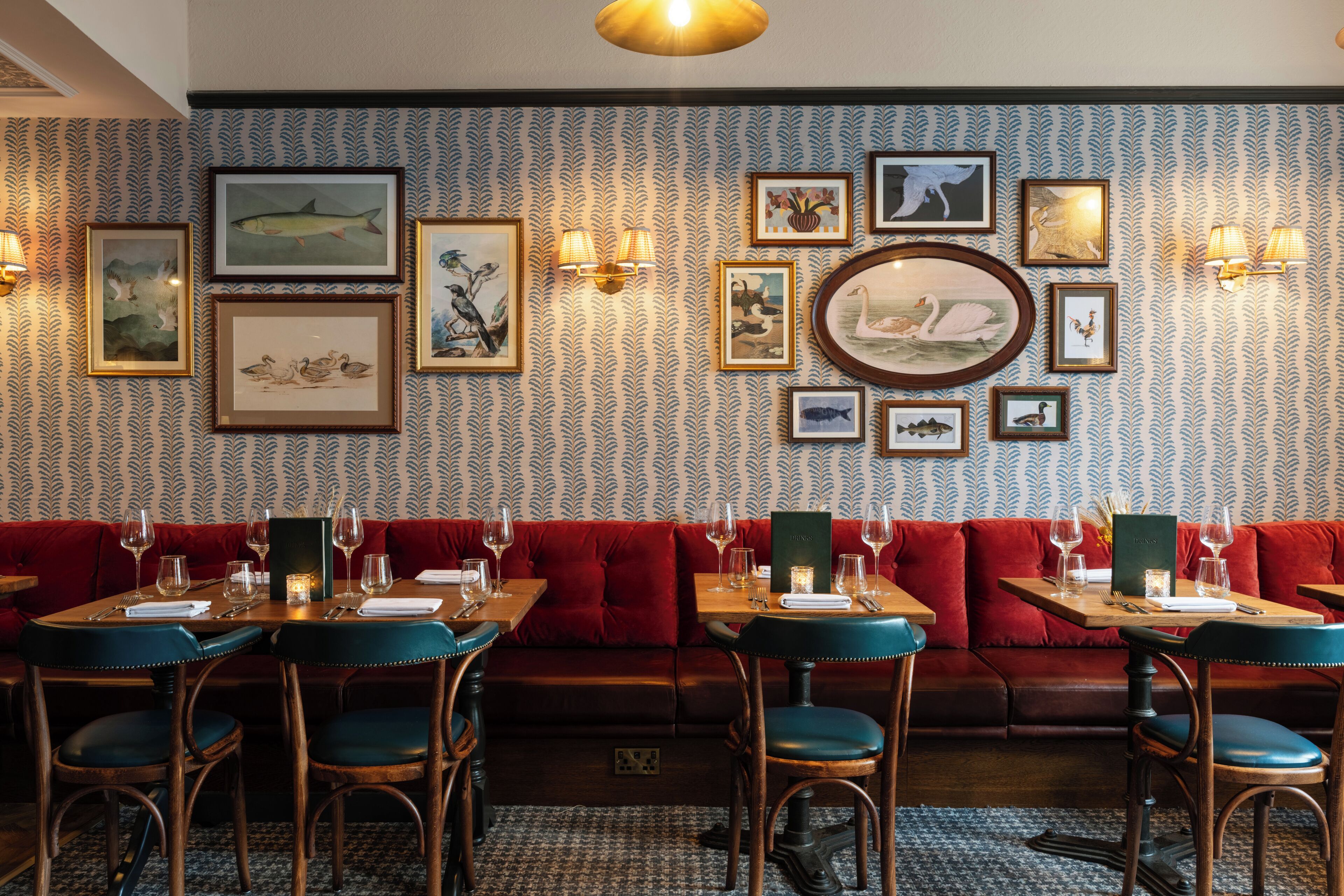 Restaurante com papel de parede estampado, obras de arte emolduradas, assentos acolchoados vermelhos e mesas de madeira.