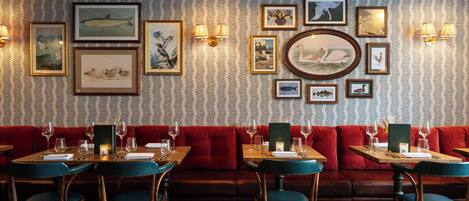 Restaurante com papel de parede estampado, obras de arte emolduradas, assentos acolchoados vermelhos e mesas de madeira.