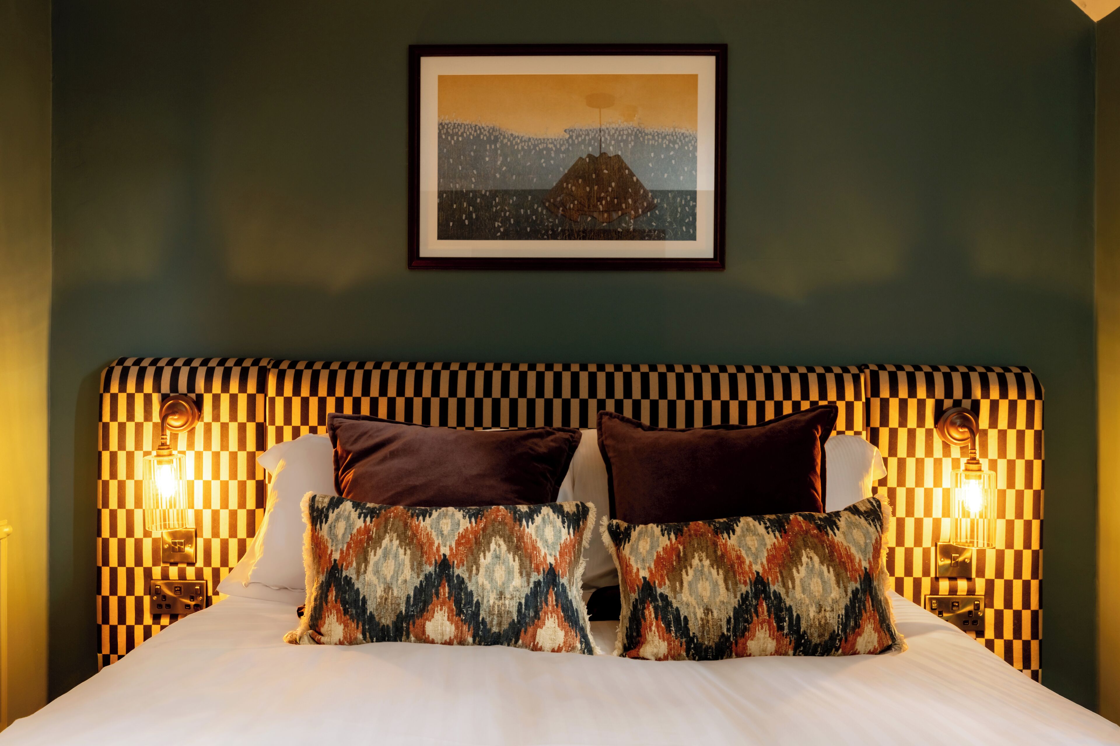A hotel room with a checkered headboard, two patterned pillows, a framed picture on the wall, and wall-mounted lamps.