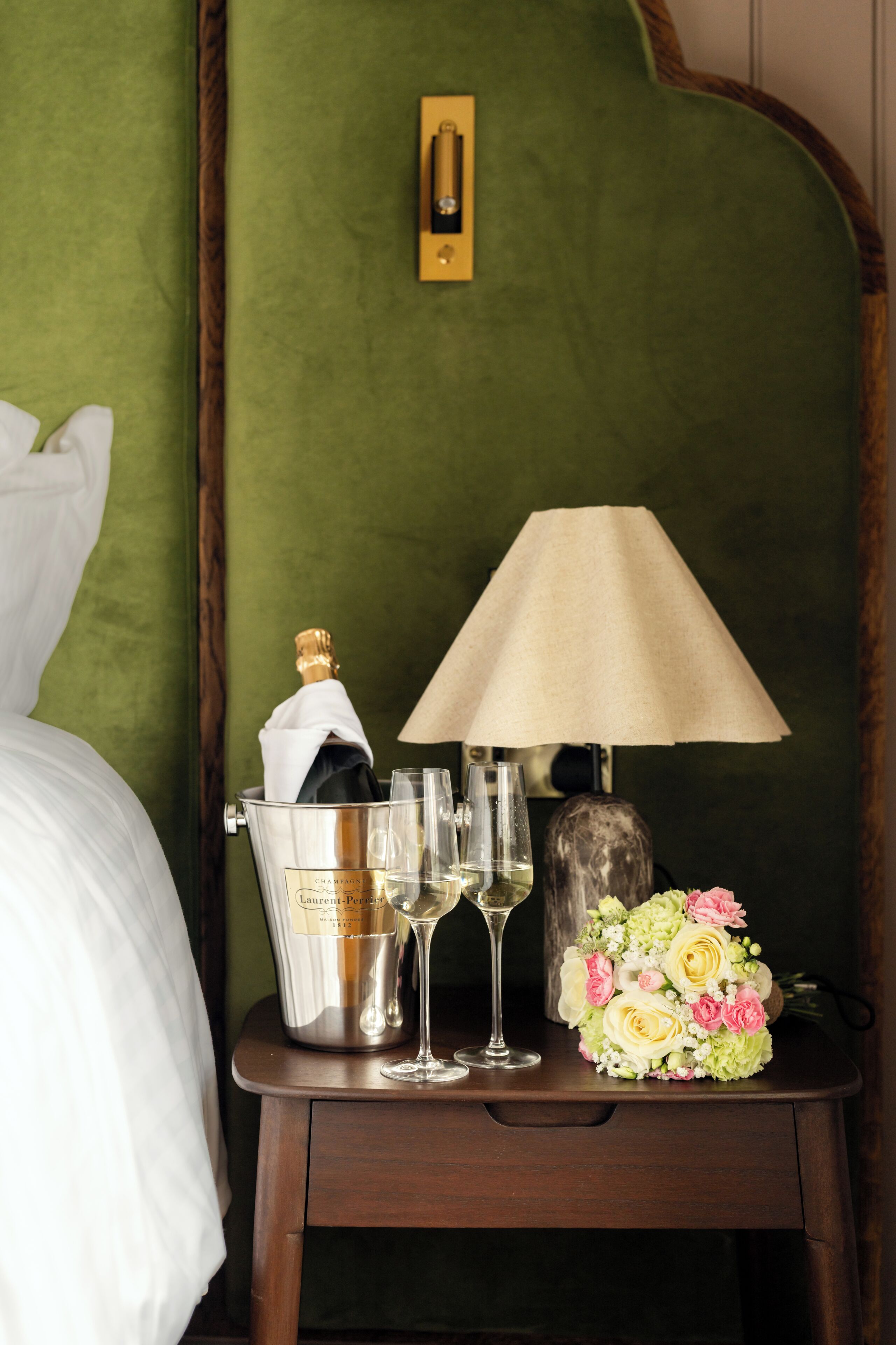 A bed, a bedside table with a bottle of champagne, two wine glasses, a lamp, and a bouquet of flowers.
