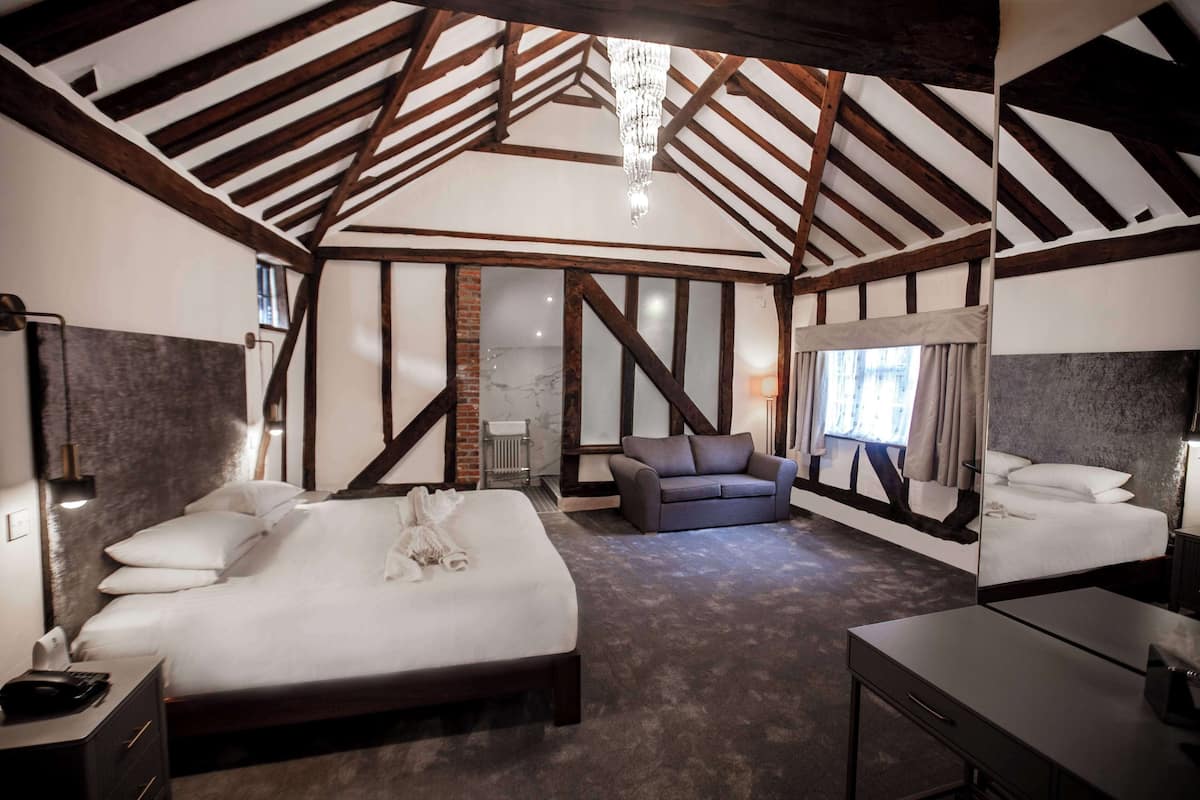 A spacious bedroom with a large bed, a sofa, and a desk. The room features exposed wooden beams and a chandelier.
