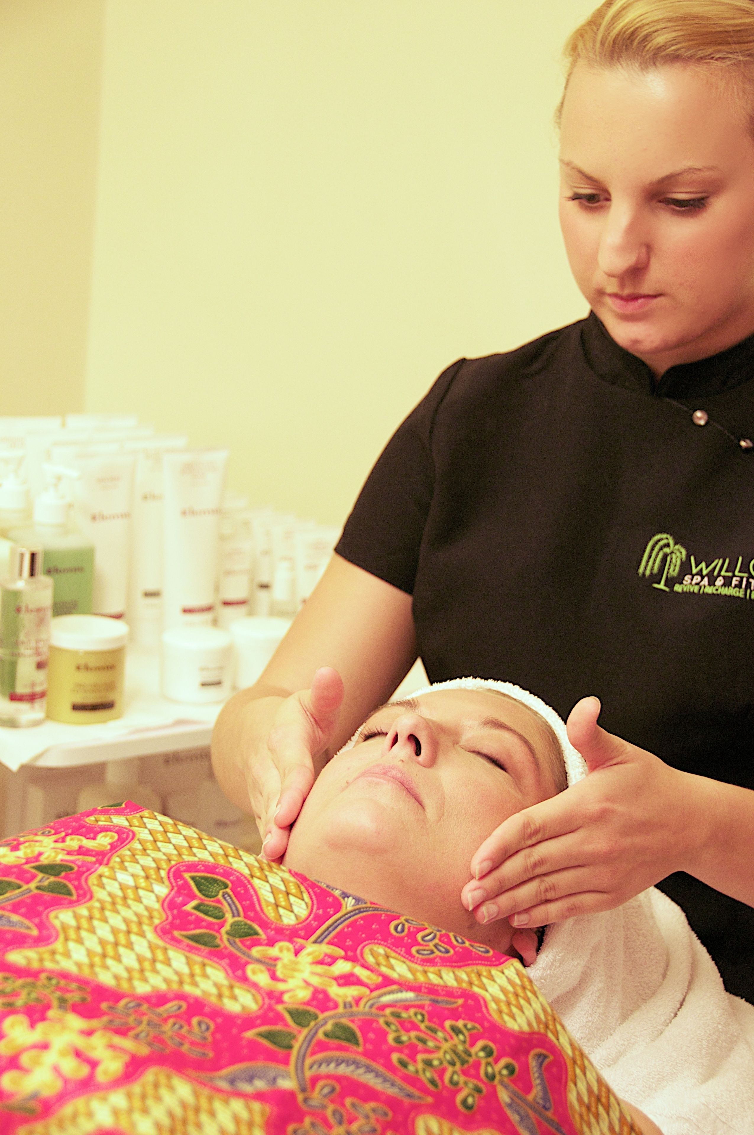 Una persona recibiendo un tratamiento facial en un spa, con varios productos para el cuidado de la piel visibles al fondo.