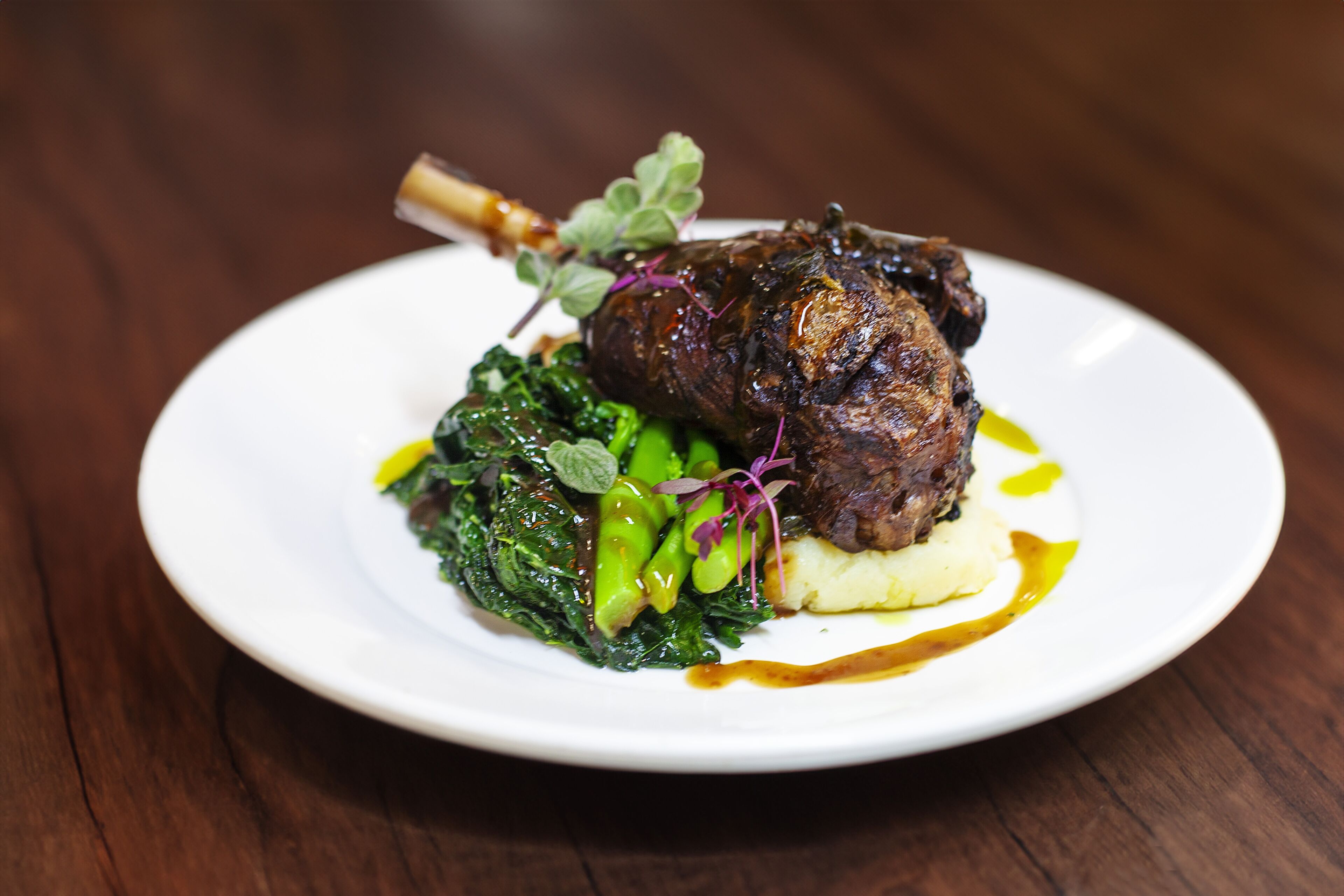 A plate of food with a lamb shank, greens, and mashed potatoes.