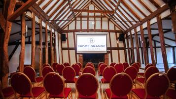 Een congreszaal met rijen rode stoelen en een groot scherm waarop 'BROME GRANGE HOTEL & SPA' staat.