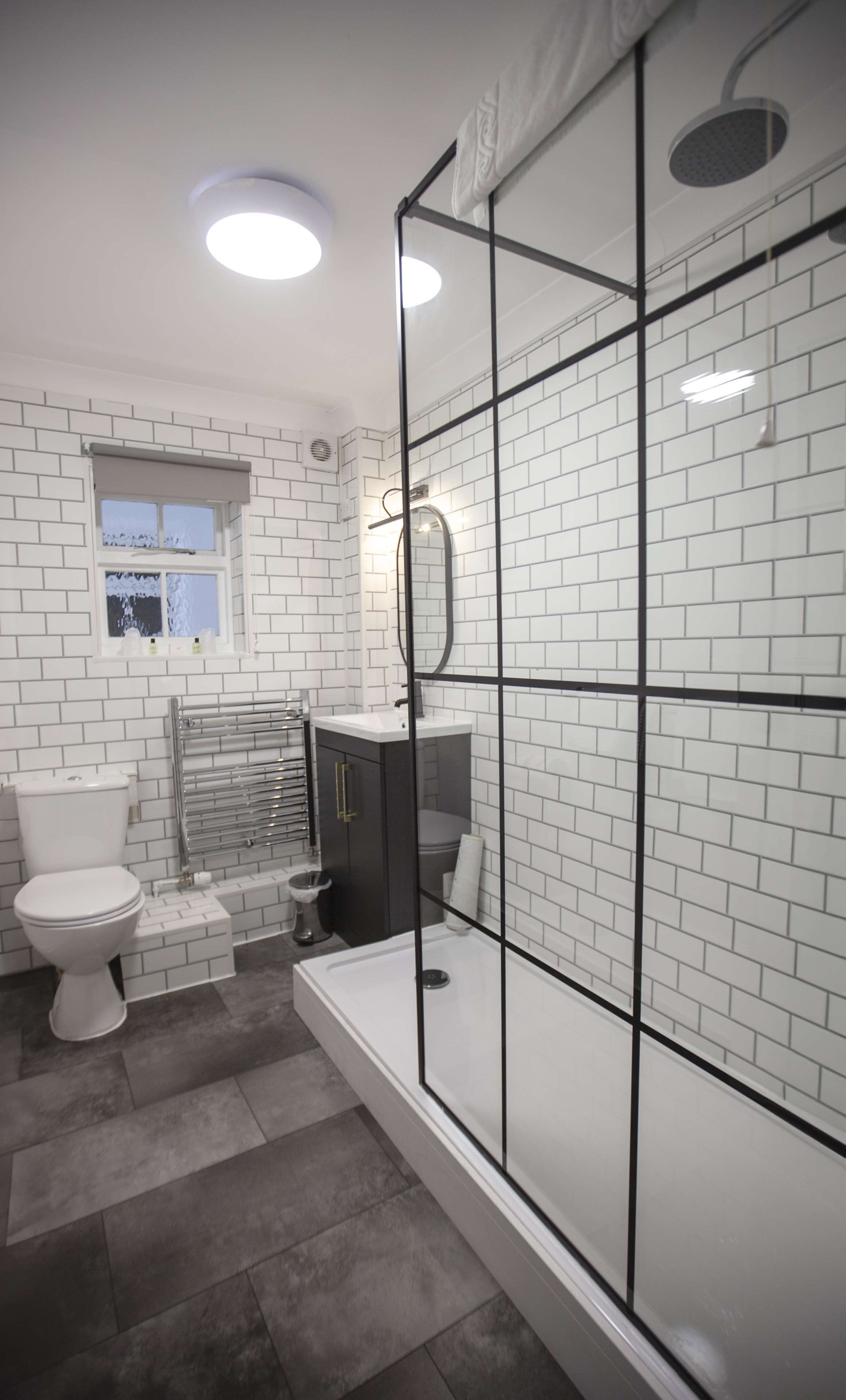 Ein modernes Badezimmer mit weißen Fliesen im U-Bahn-Stil, einer Duschkabine aus Glas, einer Toilette, einem Waschbecken und einem Fenster.