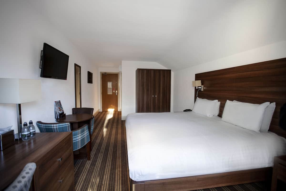 A hotel room with a large bed, a desk with a chair, a television mounted on the wall, and a wooden headboard.