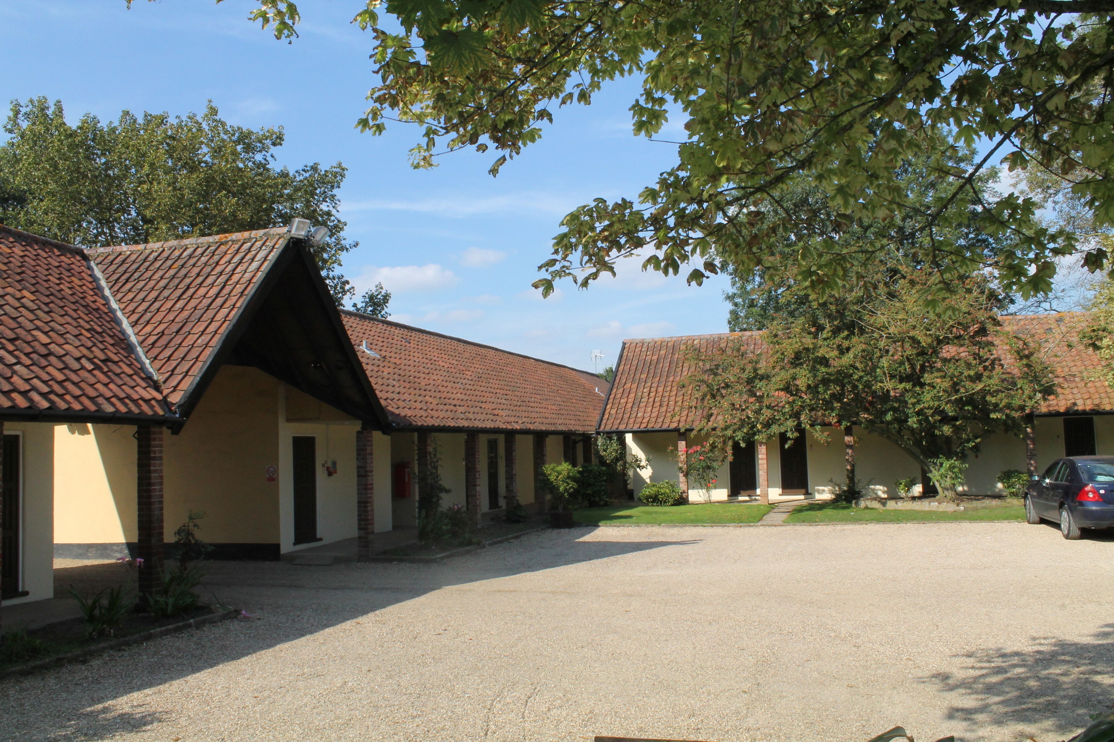 A row of single-story buildings with red-tiled roofs, a gravel driveway, and a parked car.