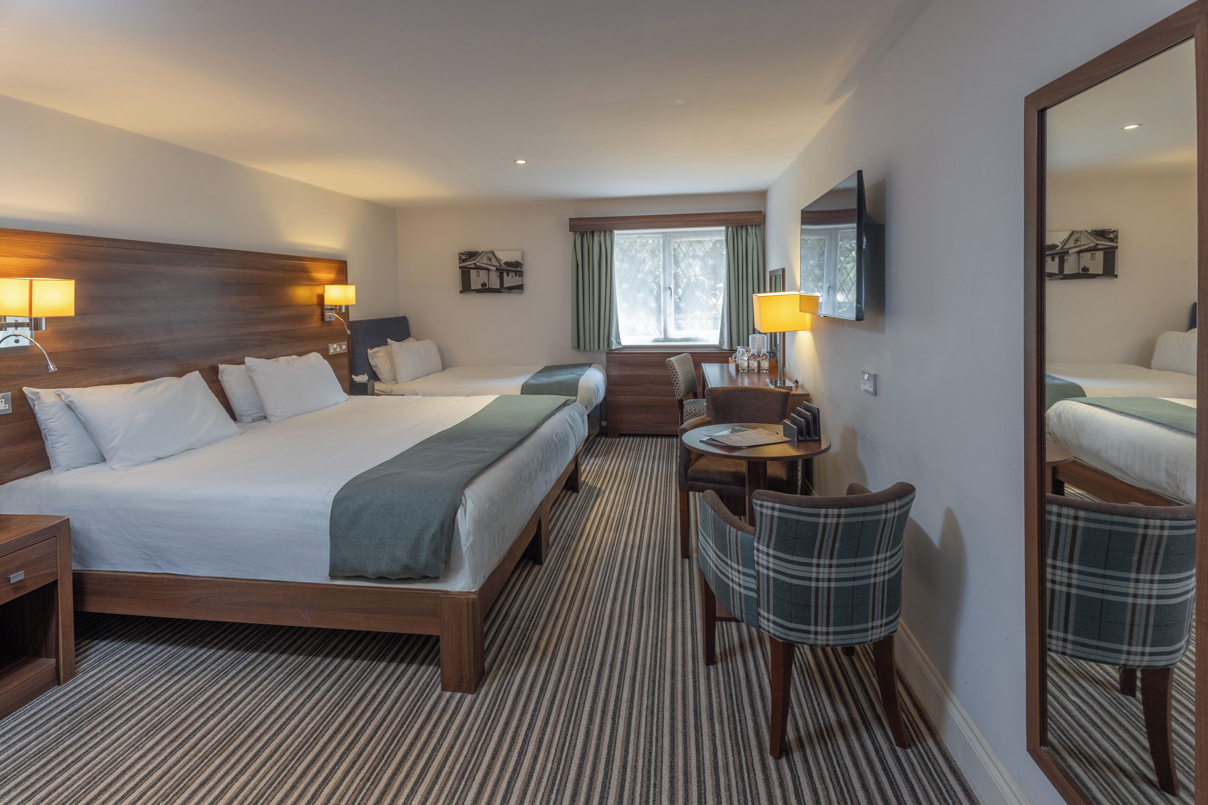 Habitación de hotel con una cama grande, un escritorio con silla, un espejo y una ventana con cortinas.