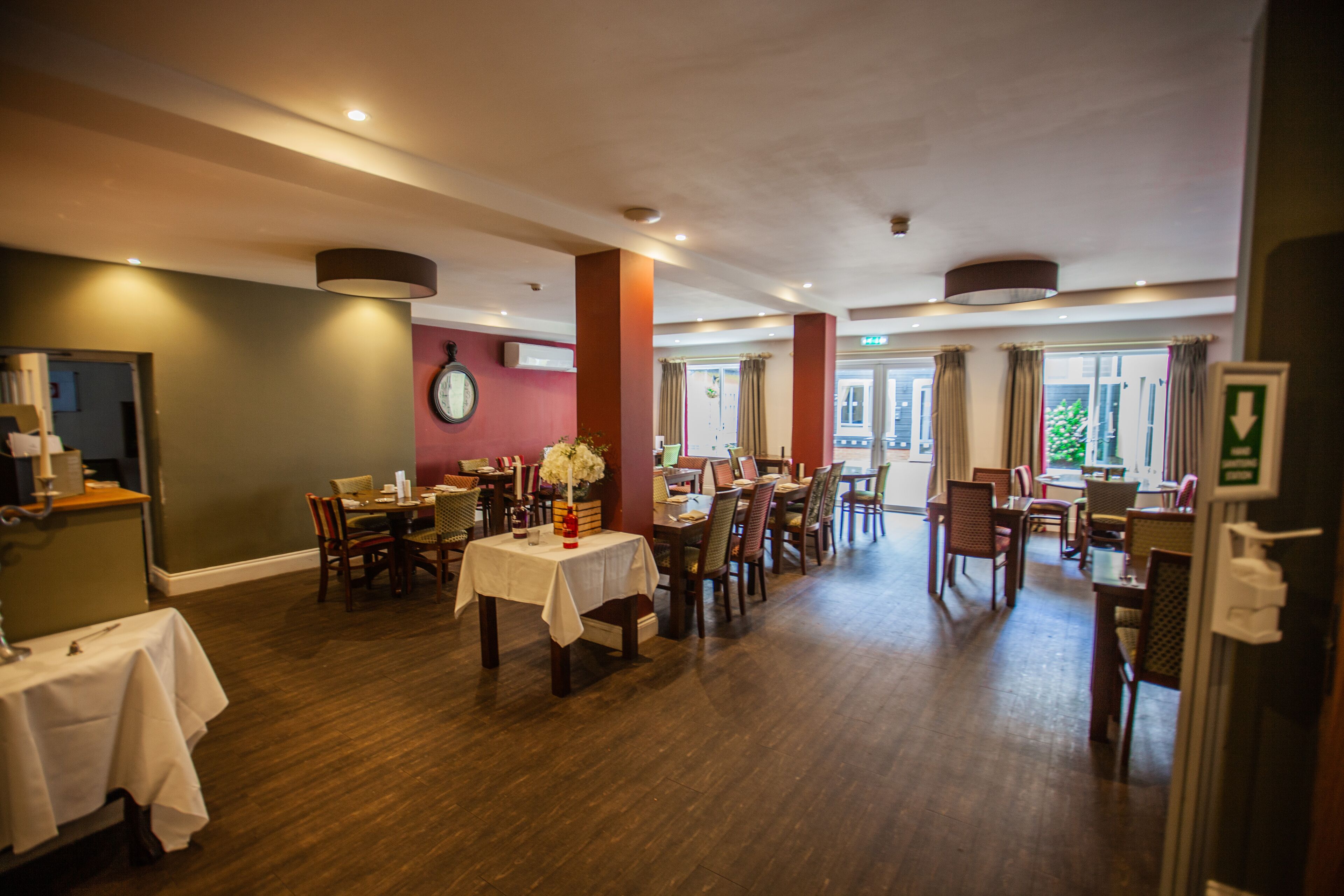 A spacious dining area with multiple tables and chairs, a clock on the wall, and large windows allowing natural light.