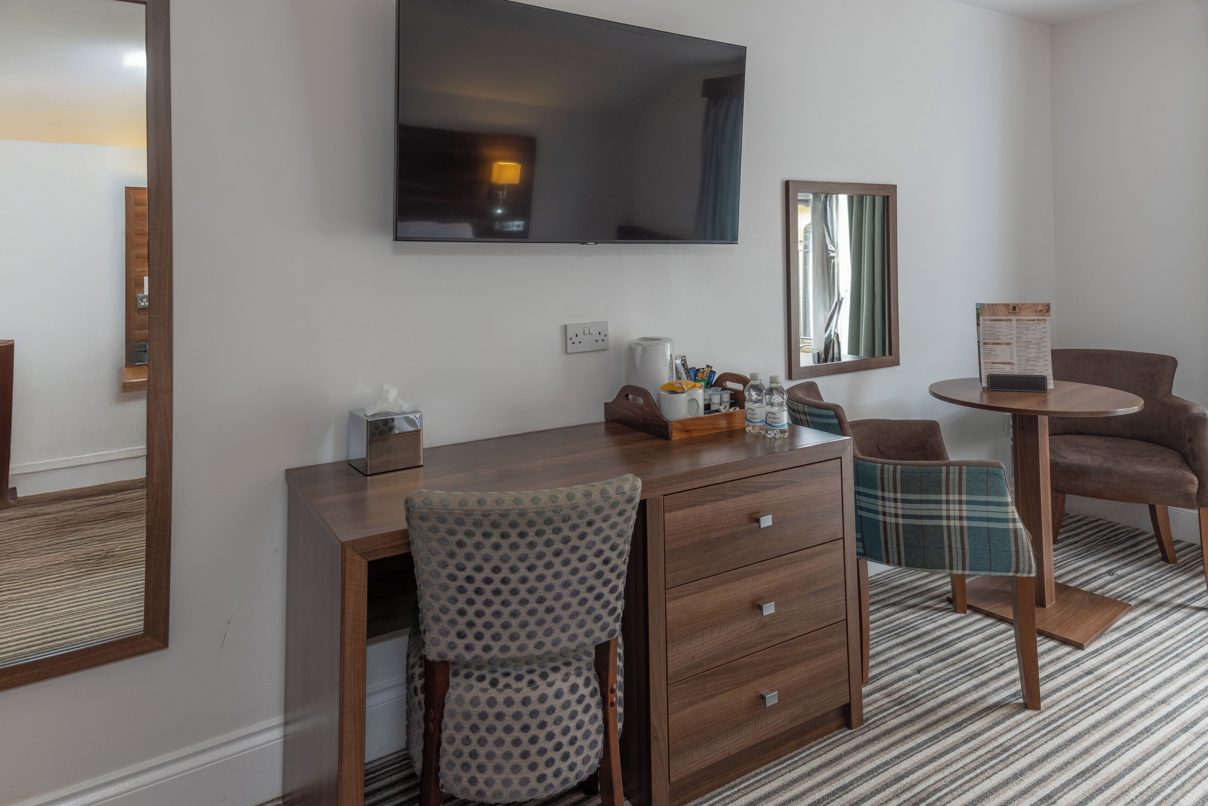 A hotel room with a wooden desk, a chair, a television, a mirror, a small table, and a patterned rug.