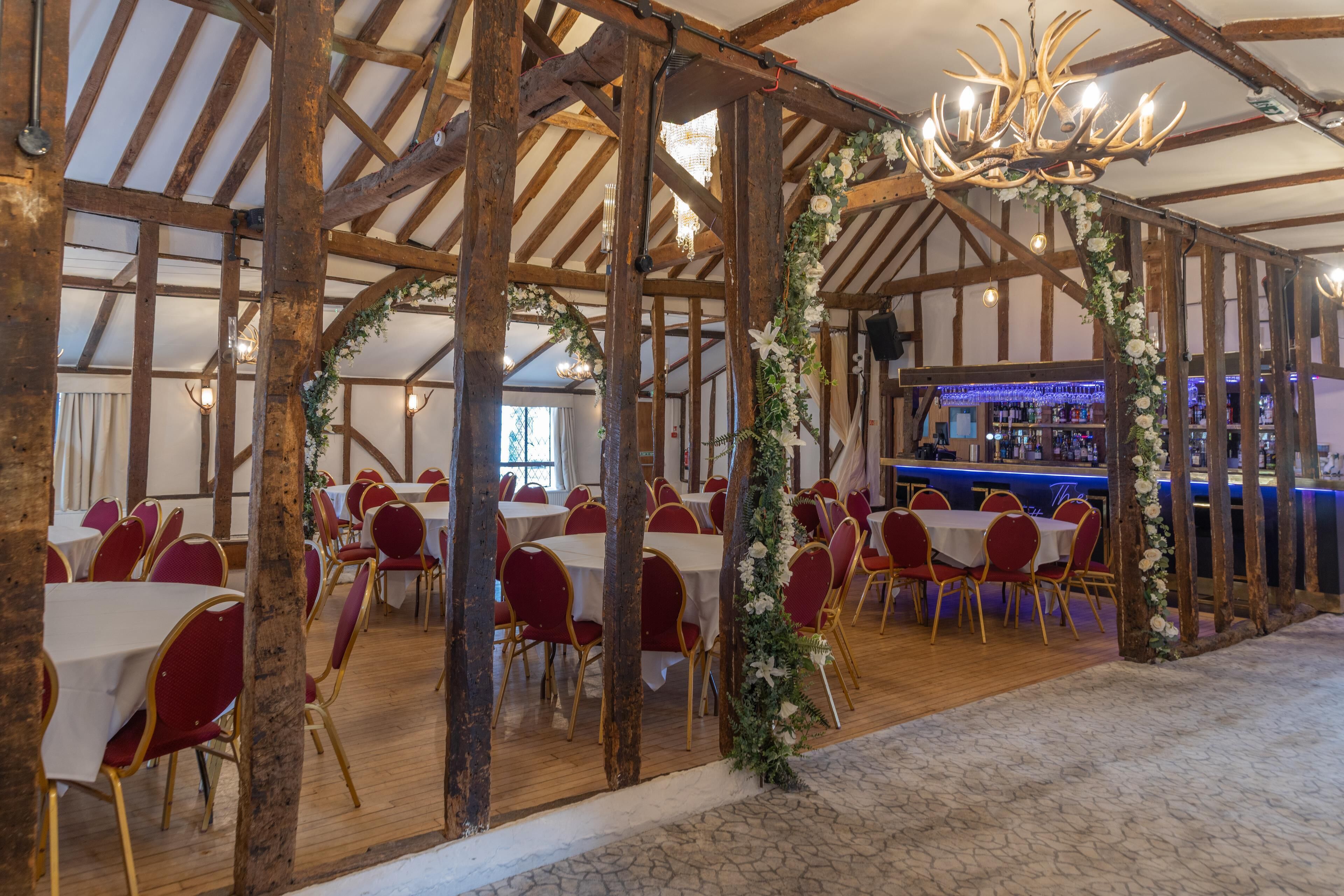 A banquet hall with wooden beams, round tables, and red chairs. A bar is visible in the background.