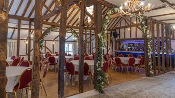 A banquet hall with wooden beams, round tables, and red chairs. A bar is visible in the background.