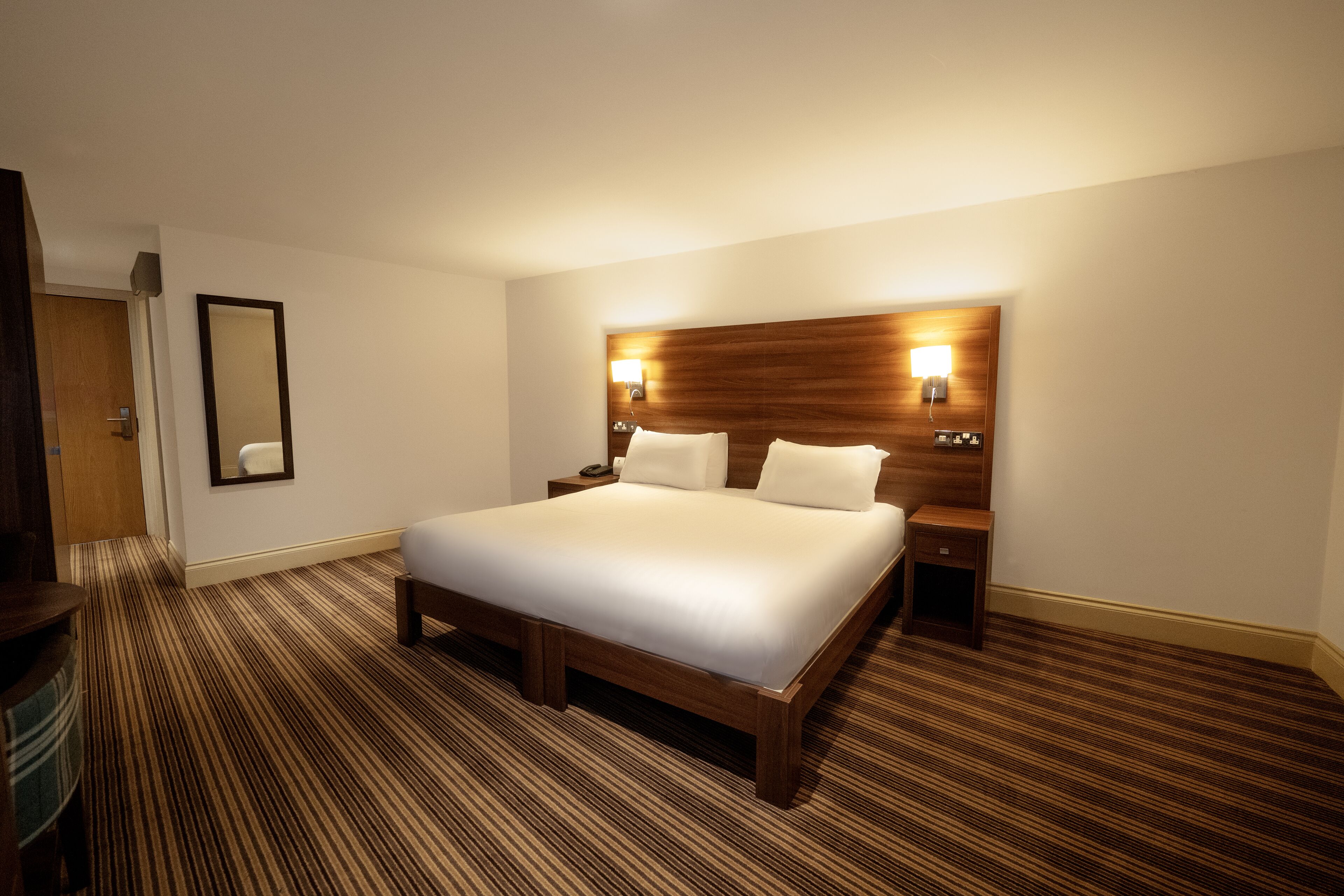 A hotel room with a large bed, wooden headboard, two bedside tables, and a carpeted floor.