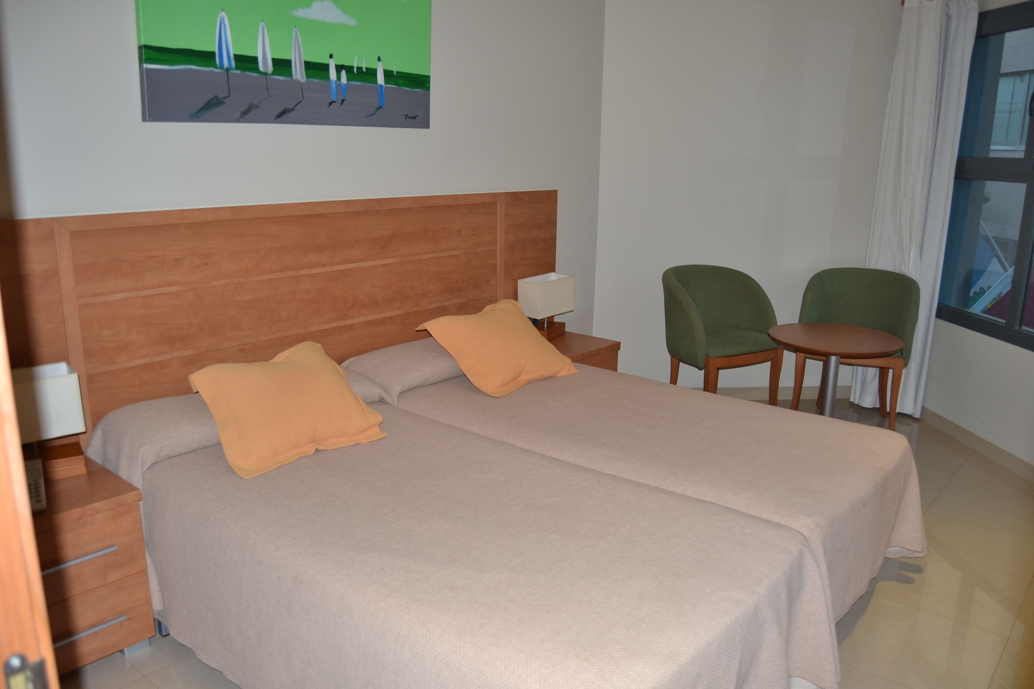 A hotel room with a large bed, two pillows, a nightstand with a lamp, two chairs, a small table, and a painting on the wall.