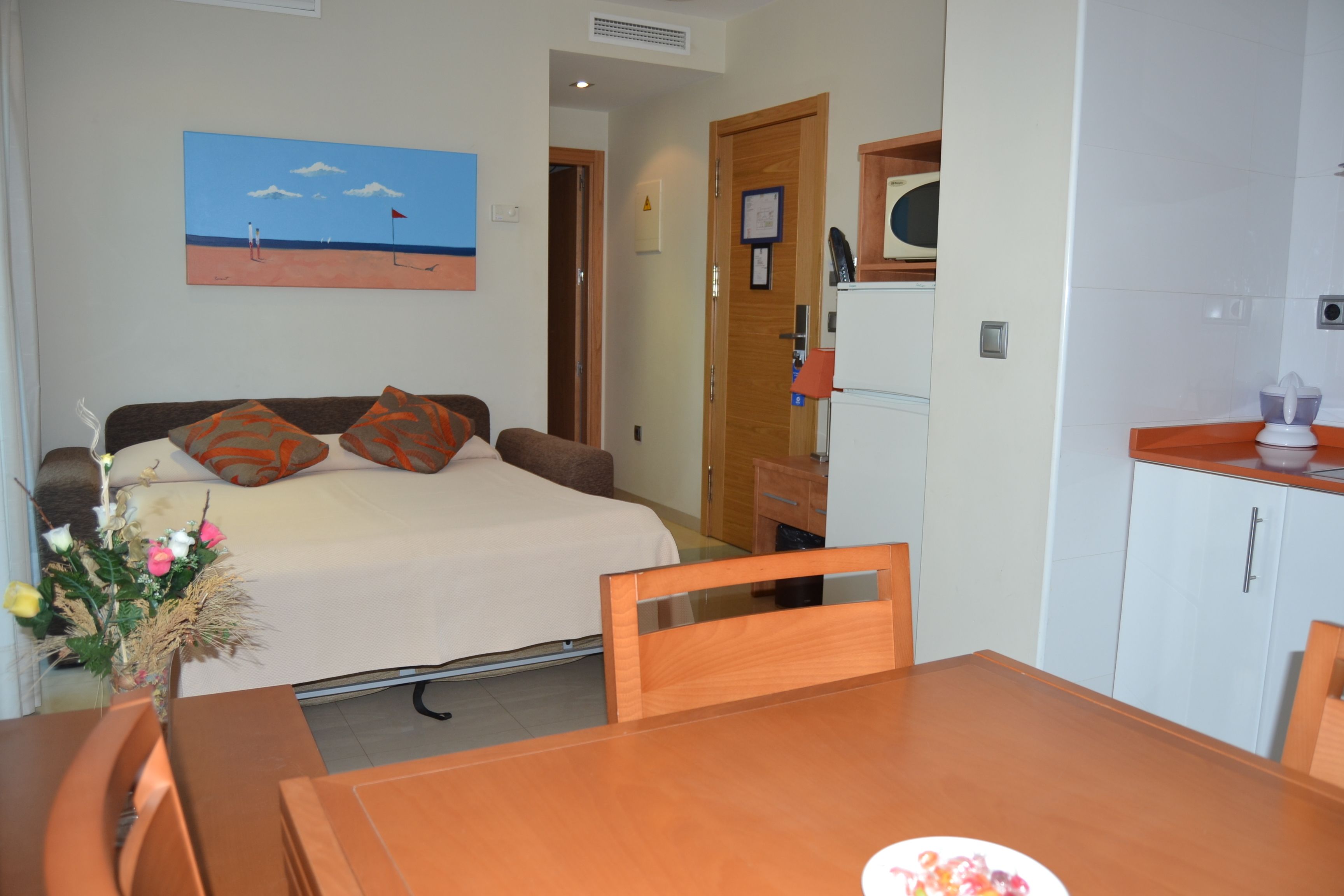 Un dormitorio con cama, cabecera de madera, una pequeña mesa con sillas, un cuadro en la pared y una zona de kitchenette.