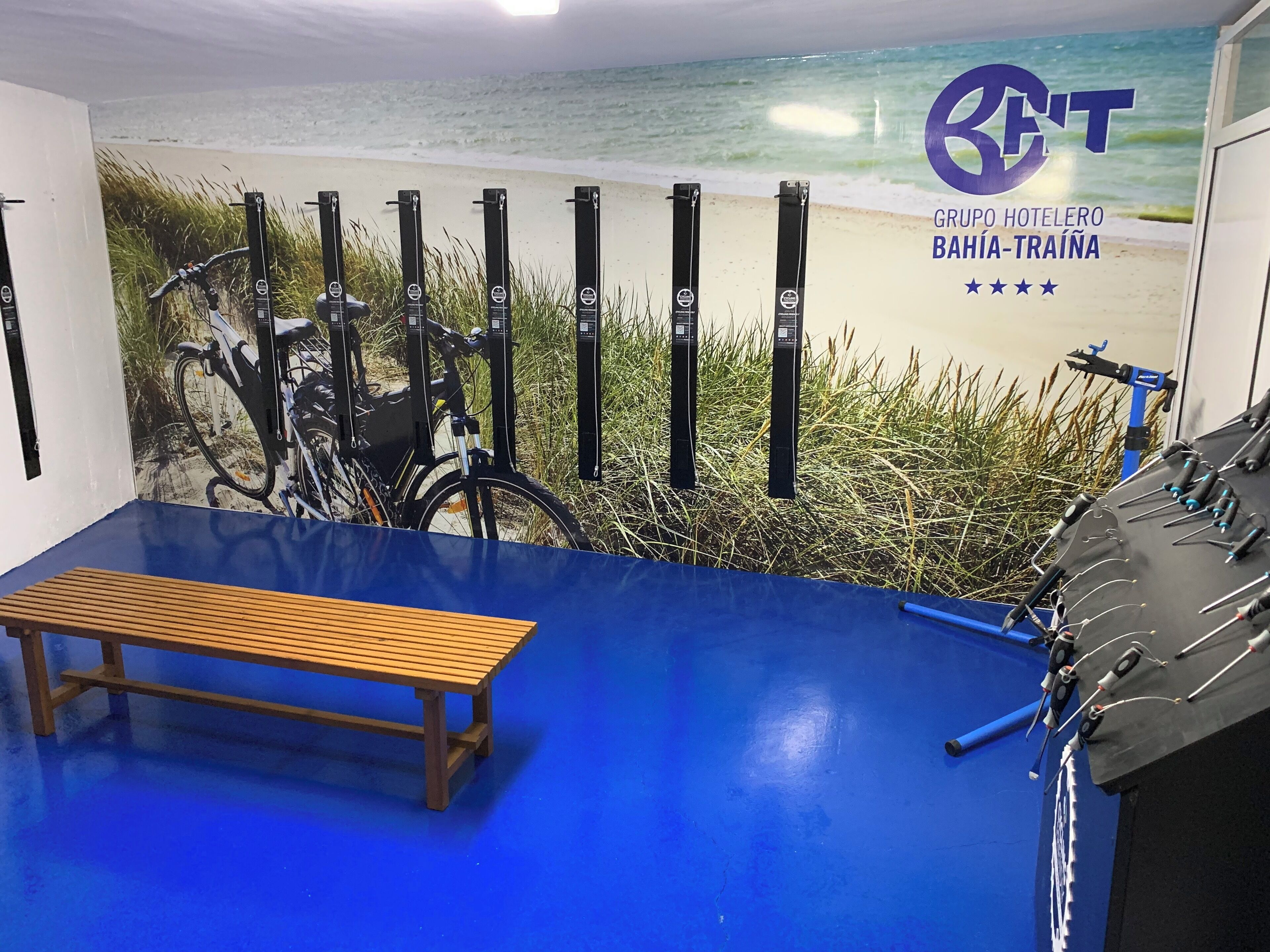 Una habitación con piso azul, un banco de madera, un mural en la pared de una playa y un soporte para bicicletas con bicicletas.