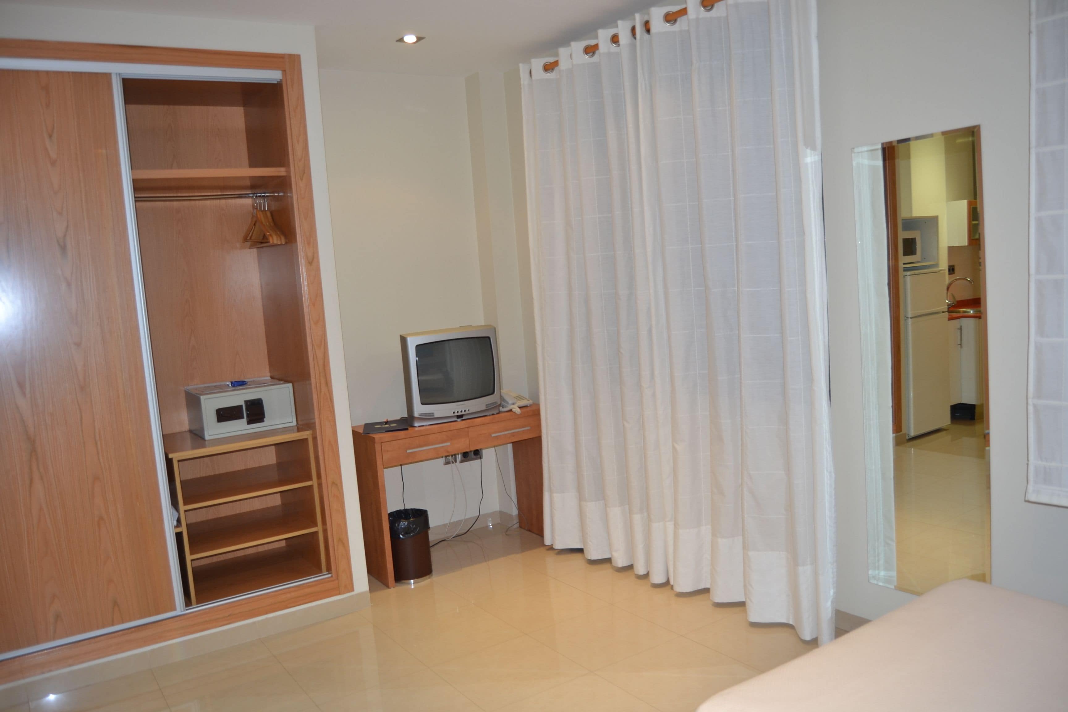 Un dormitorio con un armario de madera, un escritorio pequeño con televisor y una cortina blanca.