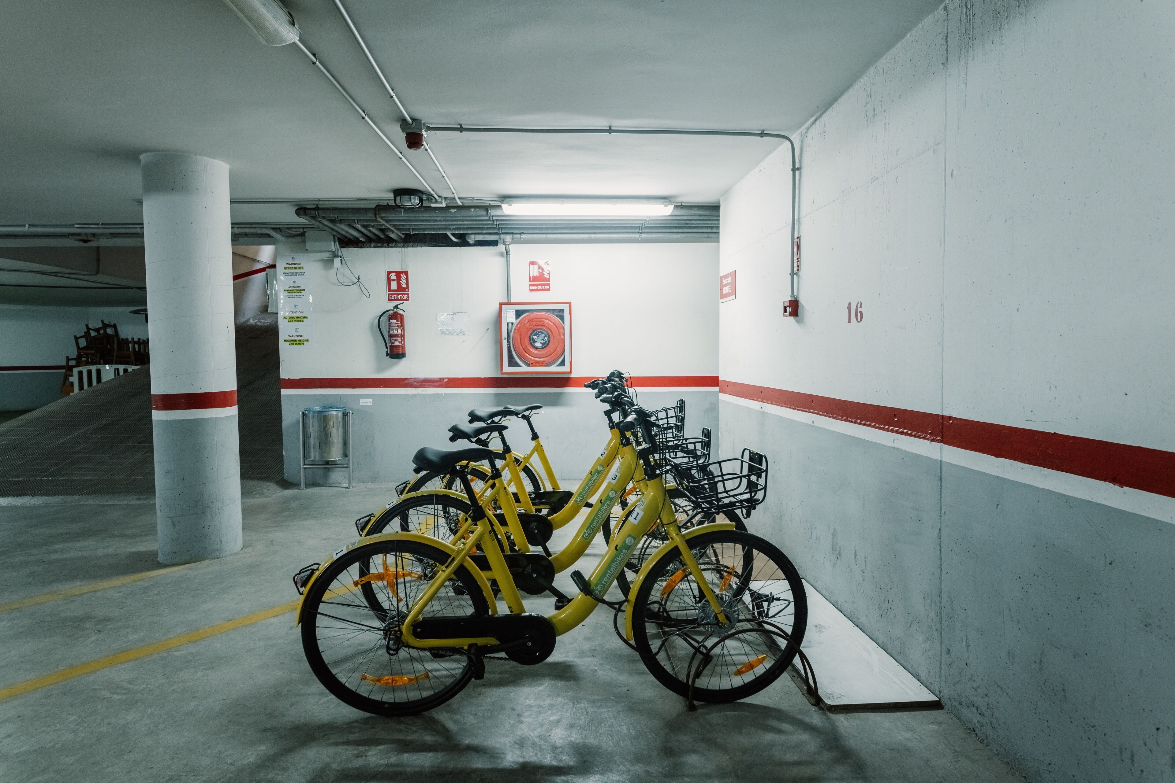 Un estacionamiento con bicicletas amarillas apoyadas en la pared, extintores y una escalera de fondo.