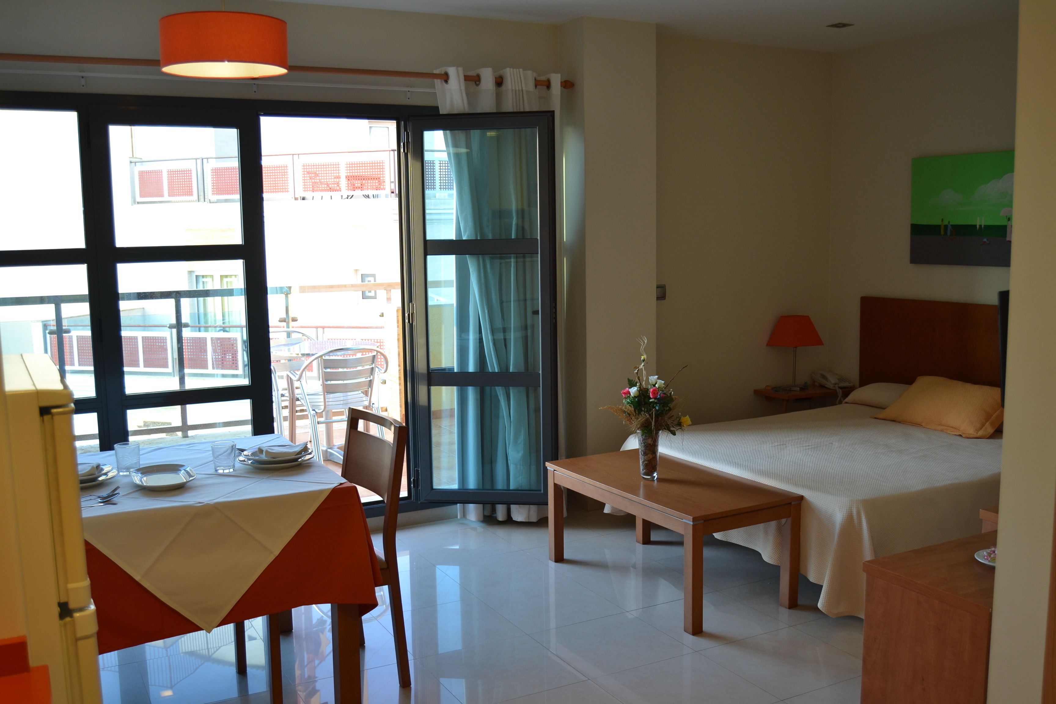 Habitación de hotel con cama, mesa, silla, escritorio, lámpara y ventana con vistas a edificios.