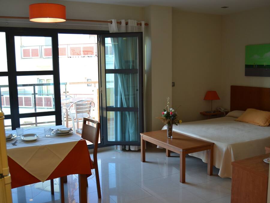 A hotel room with a bed, a table, a chair, a desk, a lamp, and a window with a view of buildings.