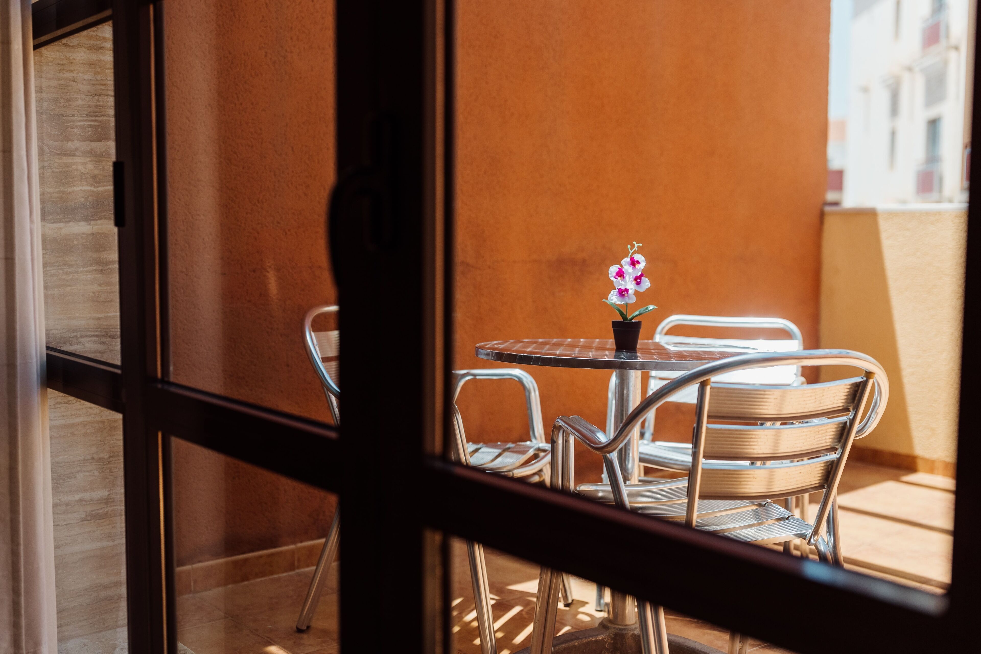 A view through a window of a dining area with a round table and metal chairs, a small potted plant, and a view of buildings in the background.