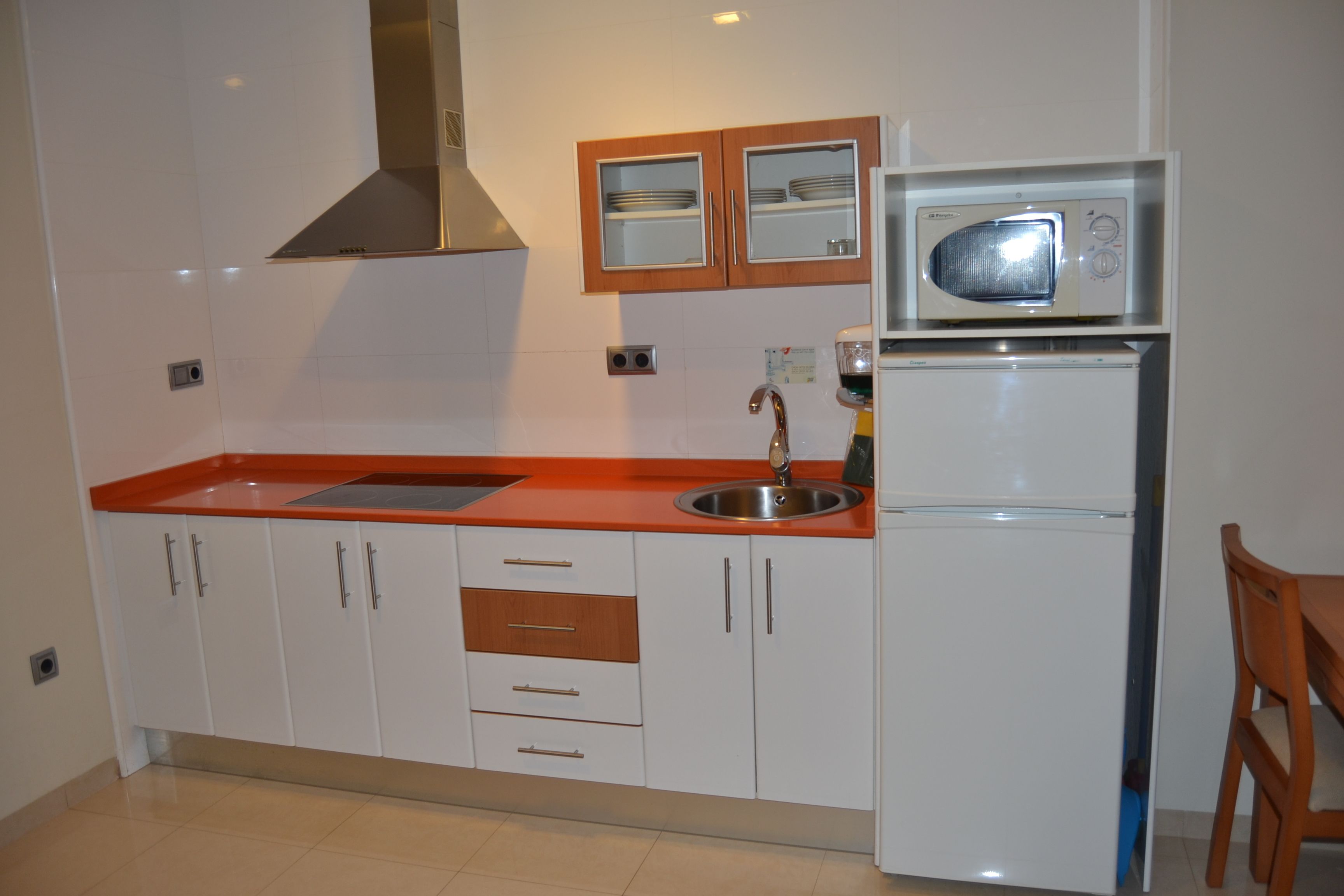 A kitchen with white cabinets, a red countertop, a stainless steel sink, a microwave, and a refrigerator.