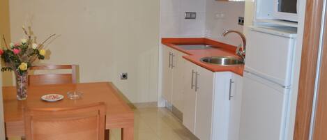 A modern kitchen with white cabinets, a stainless steel sink, and a microwave. There is a wooden dining table with chairs and a vase of flowers.