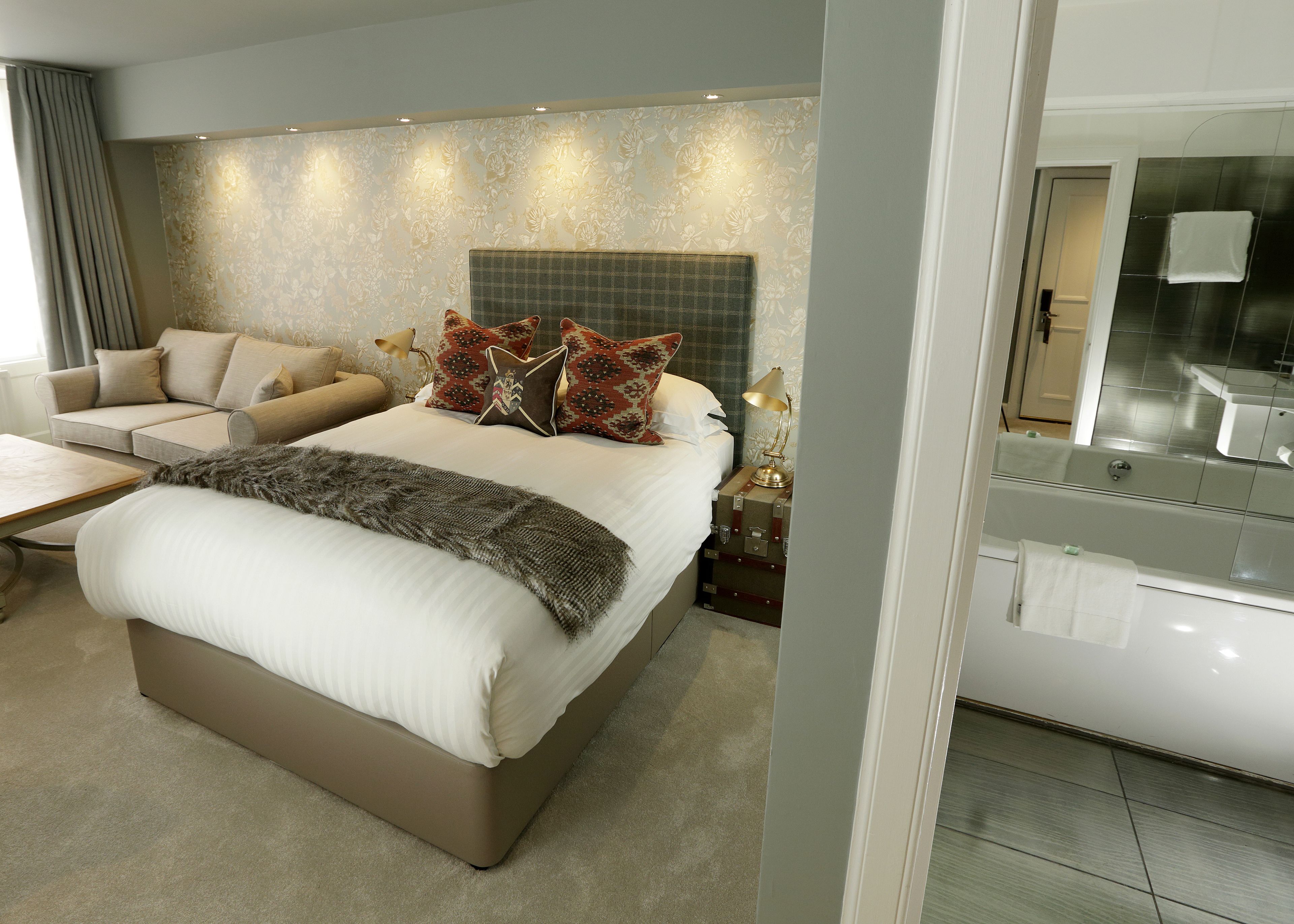A modern bedroom with a large bed, a sofa, a bedside table with a lamp, and a bathroom visible through an open door.