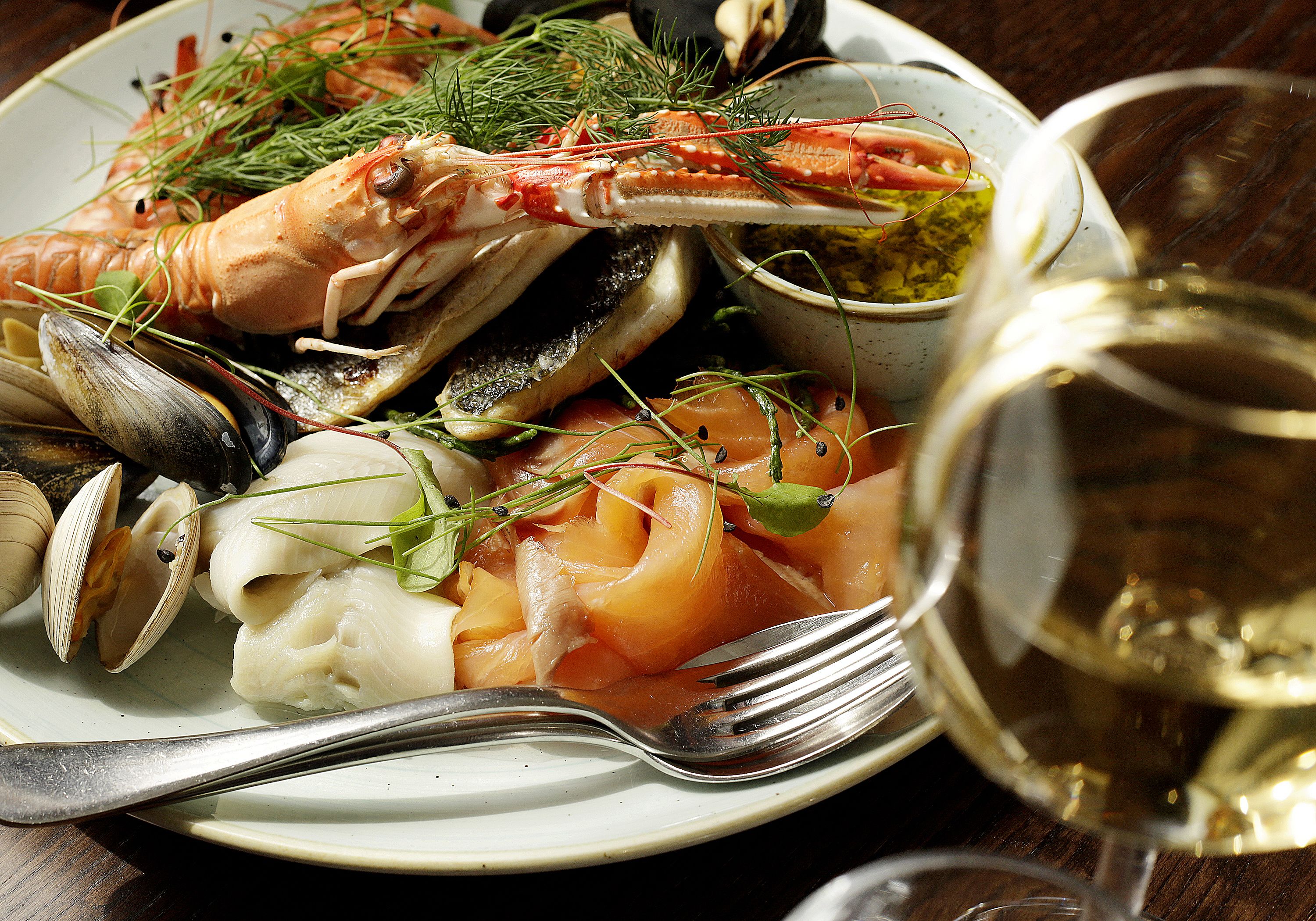 A seafood dish with mussels, clams, and shrimp, garnished with herbs, served on a plate with a fork and a glass of white wine.