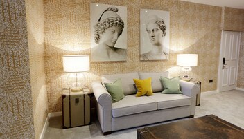 A living room with a grey sofa, green and yellow cushions, two lamps on side tables, and a patterned wallpaper.