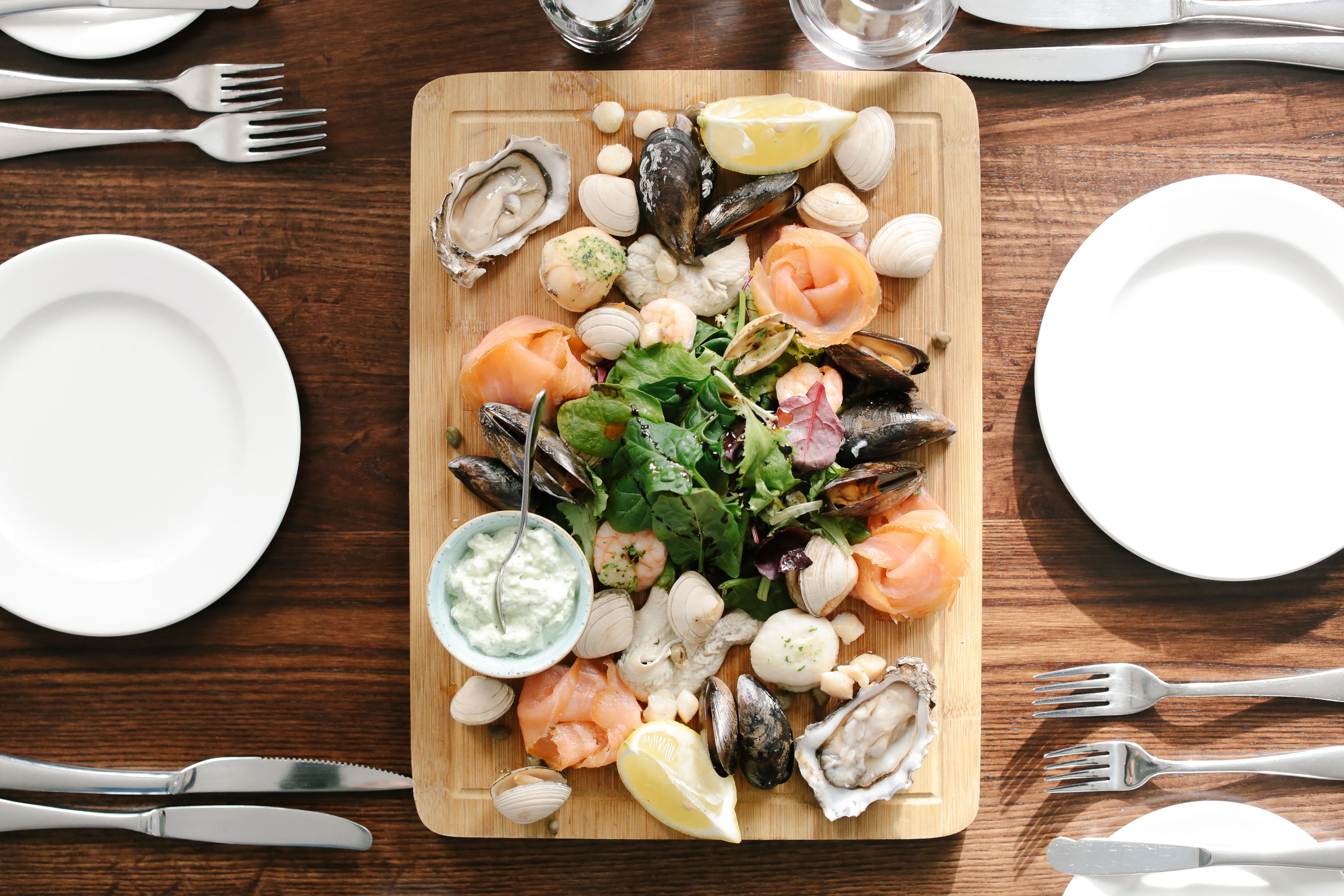 Un plateau de fruits de mer est présenté sur une planche en bois, accompagné de différentes sortes de fruits de mer, d’un quartier de citron et d’un petit bol de sauce. L’ensemble est disposé sur une table en bois, entouré d’assiettes blanches et de couverts.