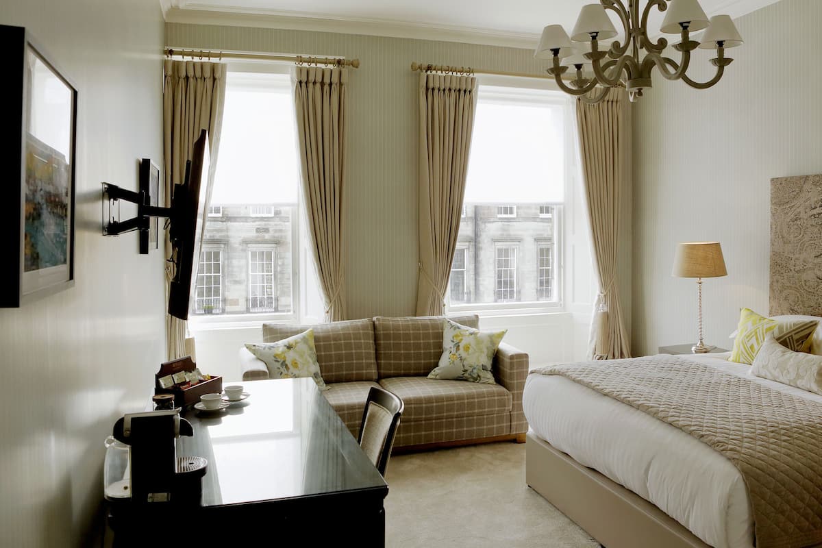 Une chambre d’hôtel avec un lit, un bureau, une chaise, une lampe et une vue sur les bâtiments par les fenêtres.